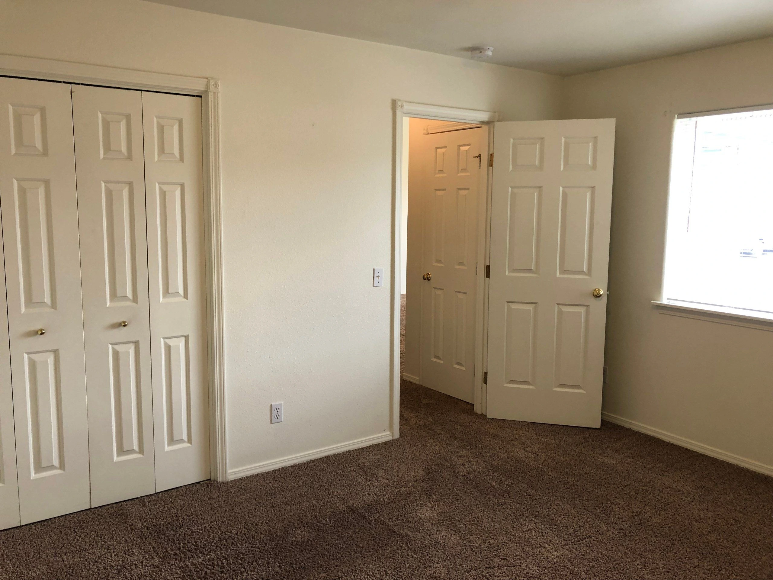 An empty carpeted room has white paneled closet doors, an open doorway, and a window with blinds.