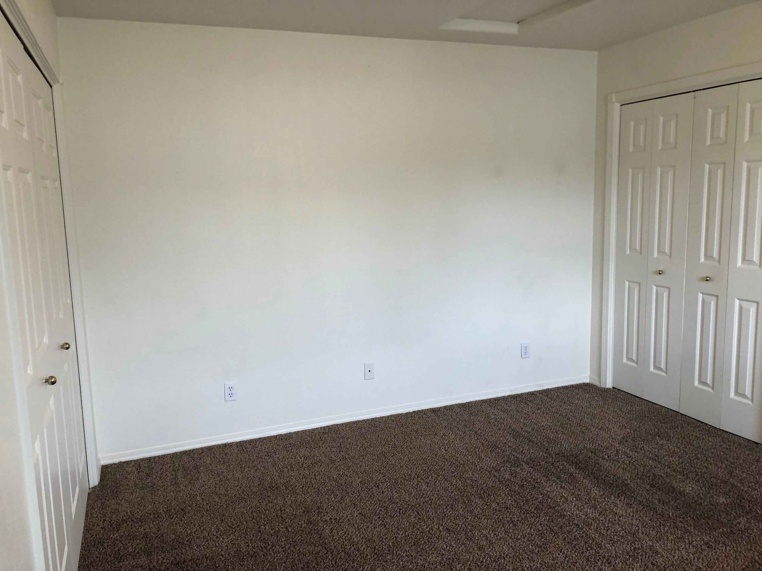 An empty room has a large white wall, brown carpet, a paneled closet, and a partially open door.