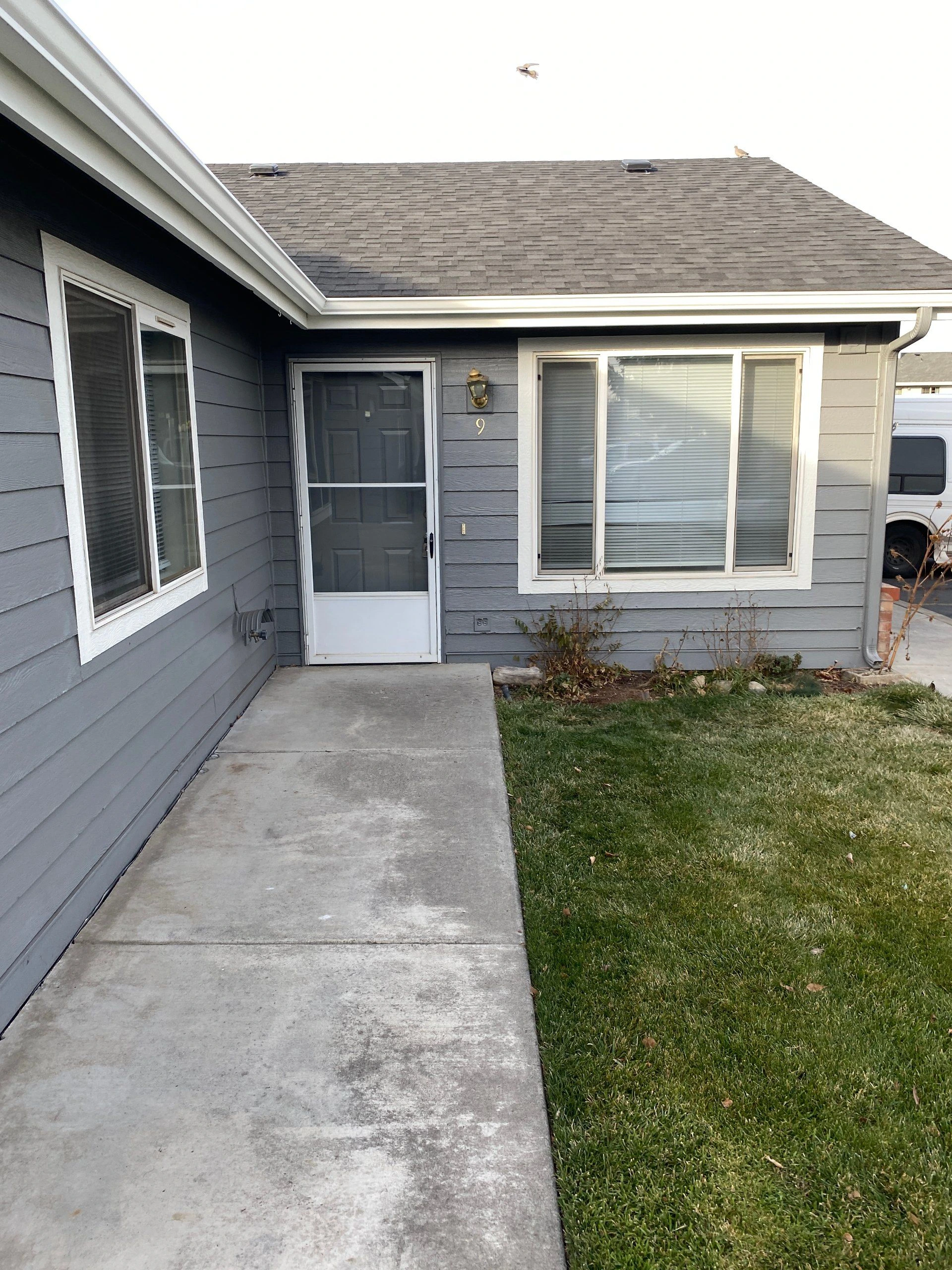 Gray-sided house exterior with a screened front door, white-trimmed windows, concrete walkway, and lawn.