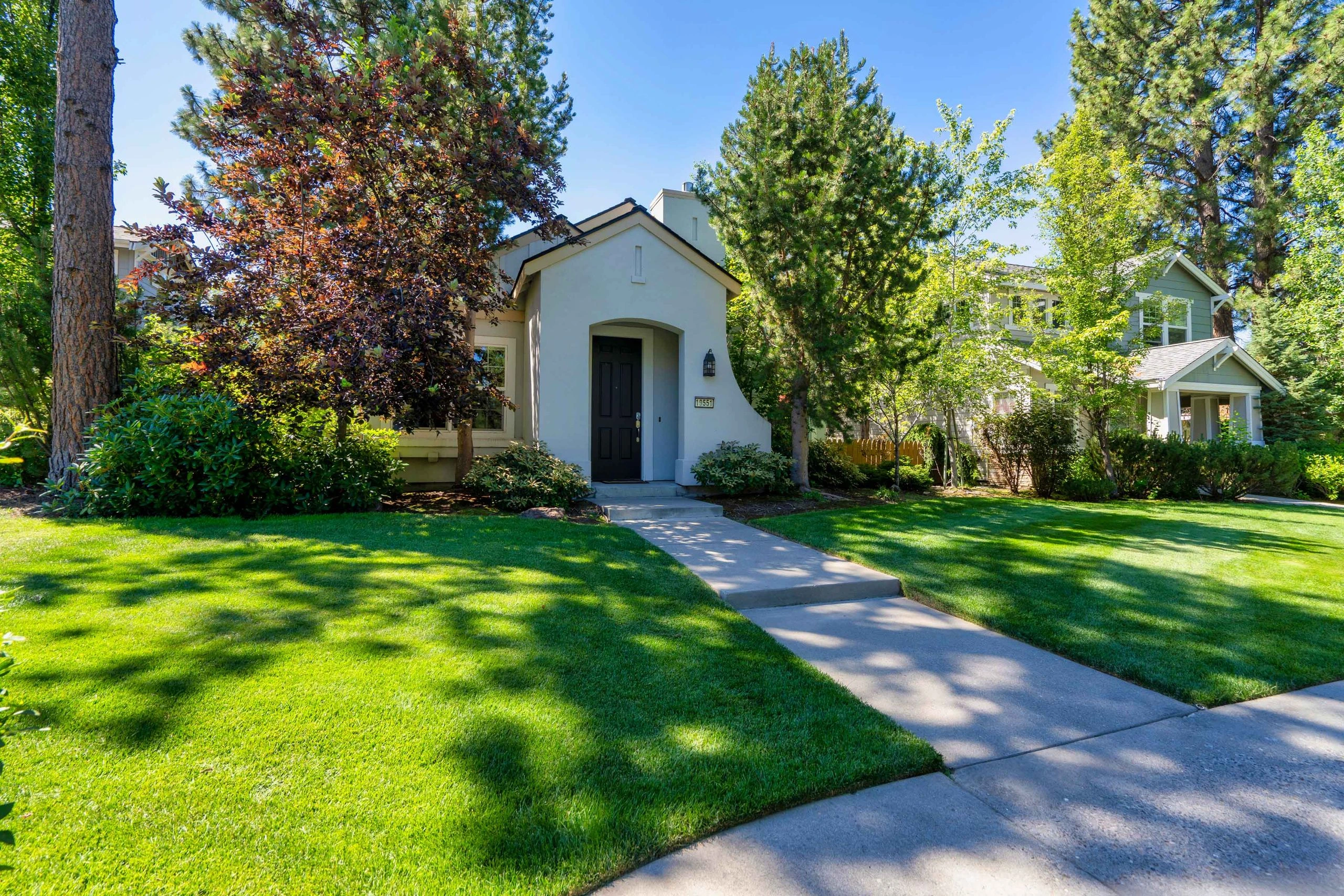 A light-colored house with a black front door stands among trees, behind a green lawn and concrete walkway.