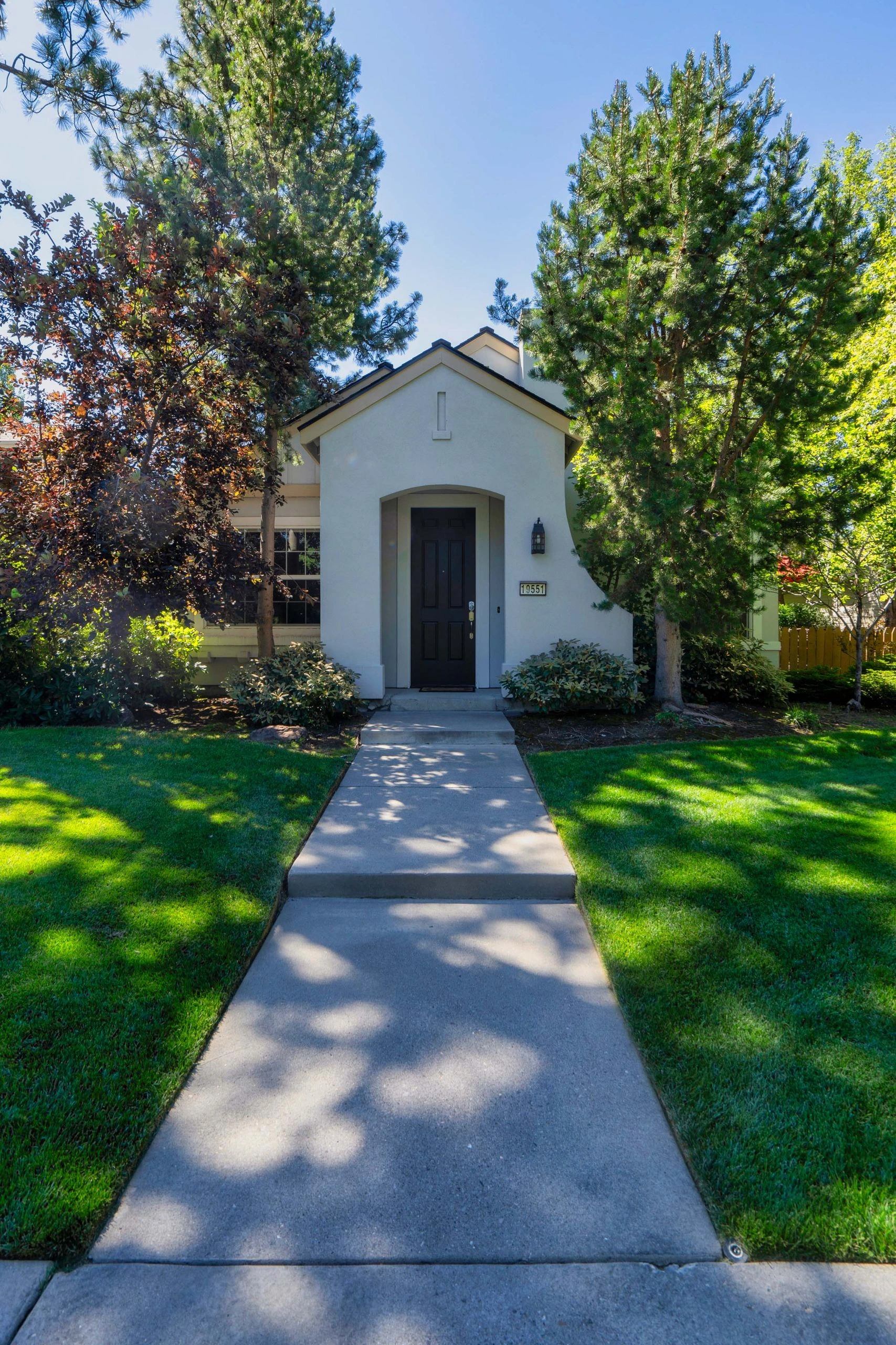 A concrete walkway leads to a dark front door on a white house, framed by trees, shrubs, and a green lawn.