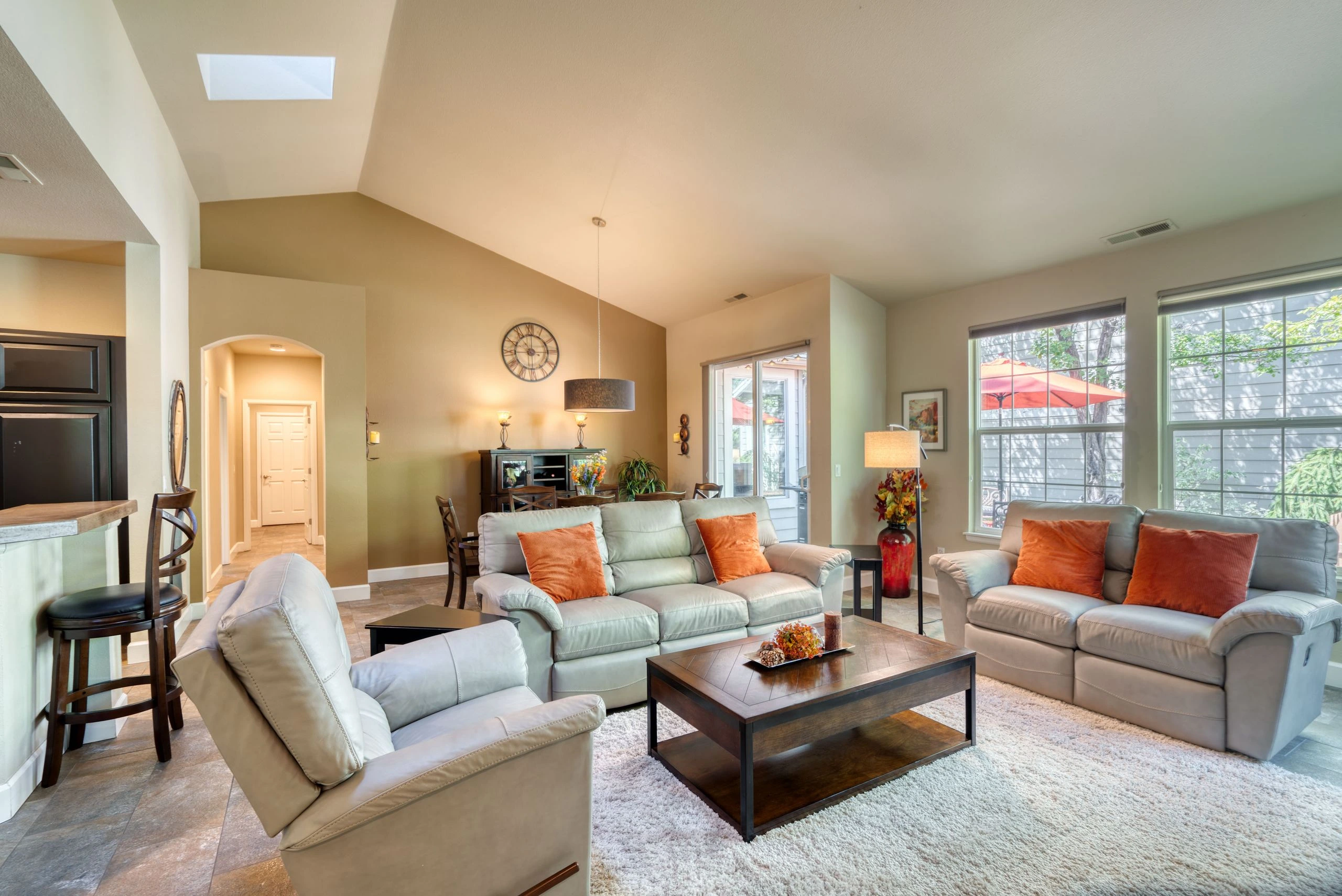 A living room with pale sofas, orange cushions, a dark coffee table, large windows, and a vaulted ceiling with a skylight.