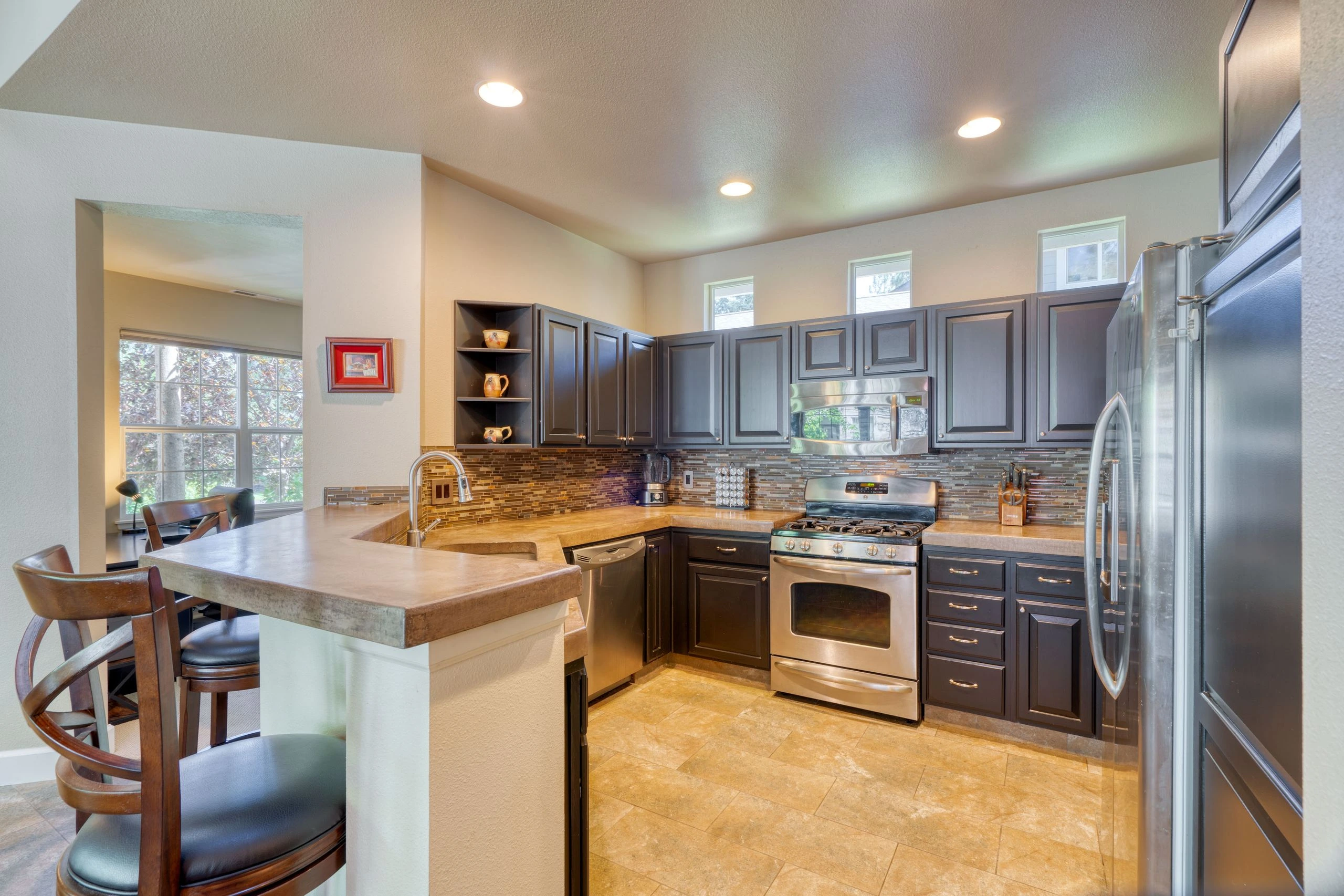 A kitchen with dark cabinets, stainless steel appliances, a tiled backsplash, and a counter with a sink and chairs.