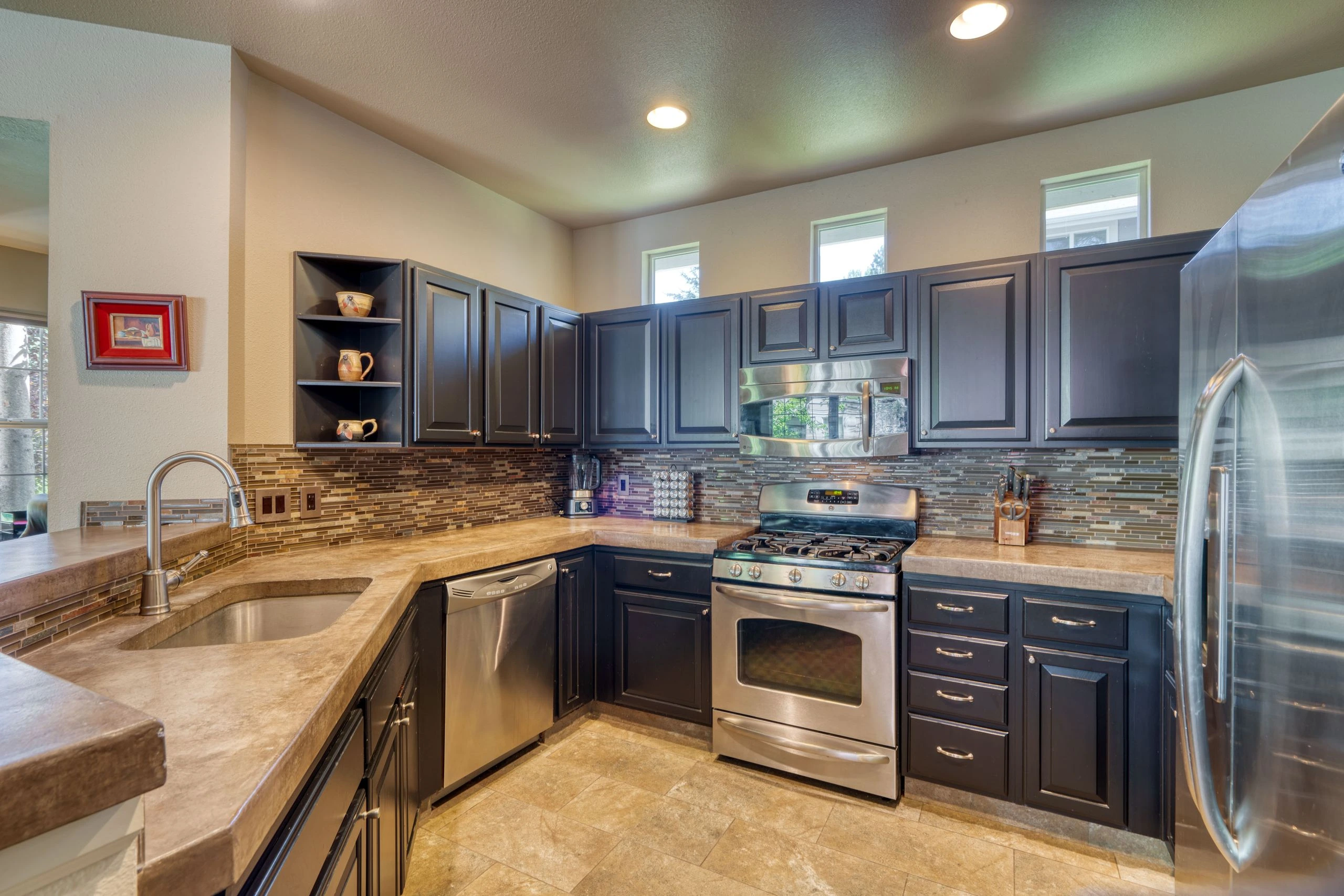 A kitchen with dark cabinets, stone counters, a tiled backsplash, a sink, and stainless steel appliances.