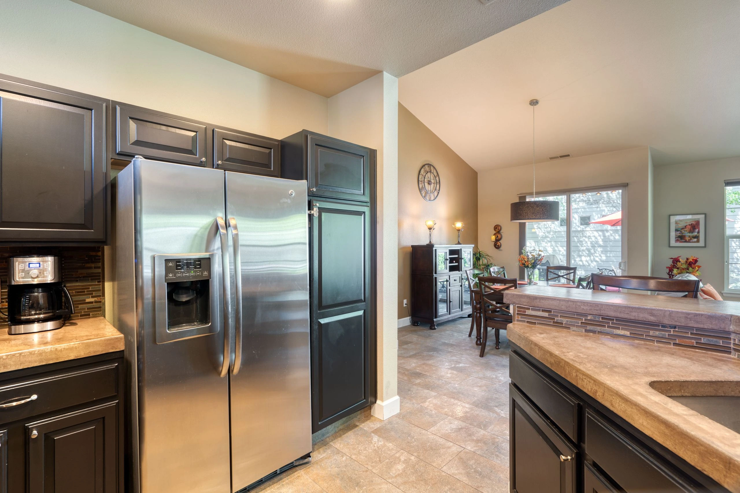 A kitchen with dark cabinets, a stainless-steel refrigerator, and a stone-topped island opens onto a dining area with a table and chairs.