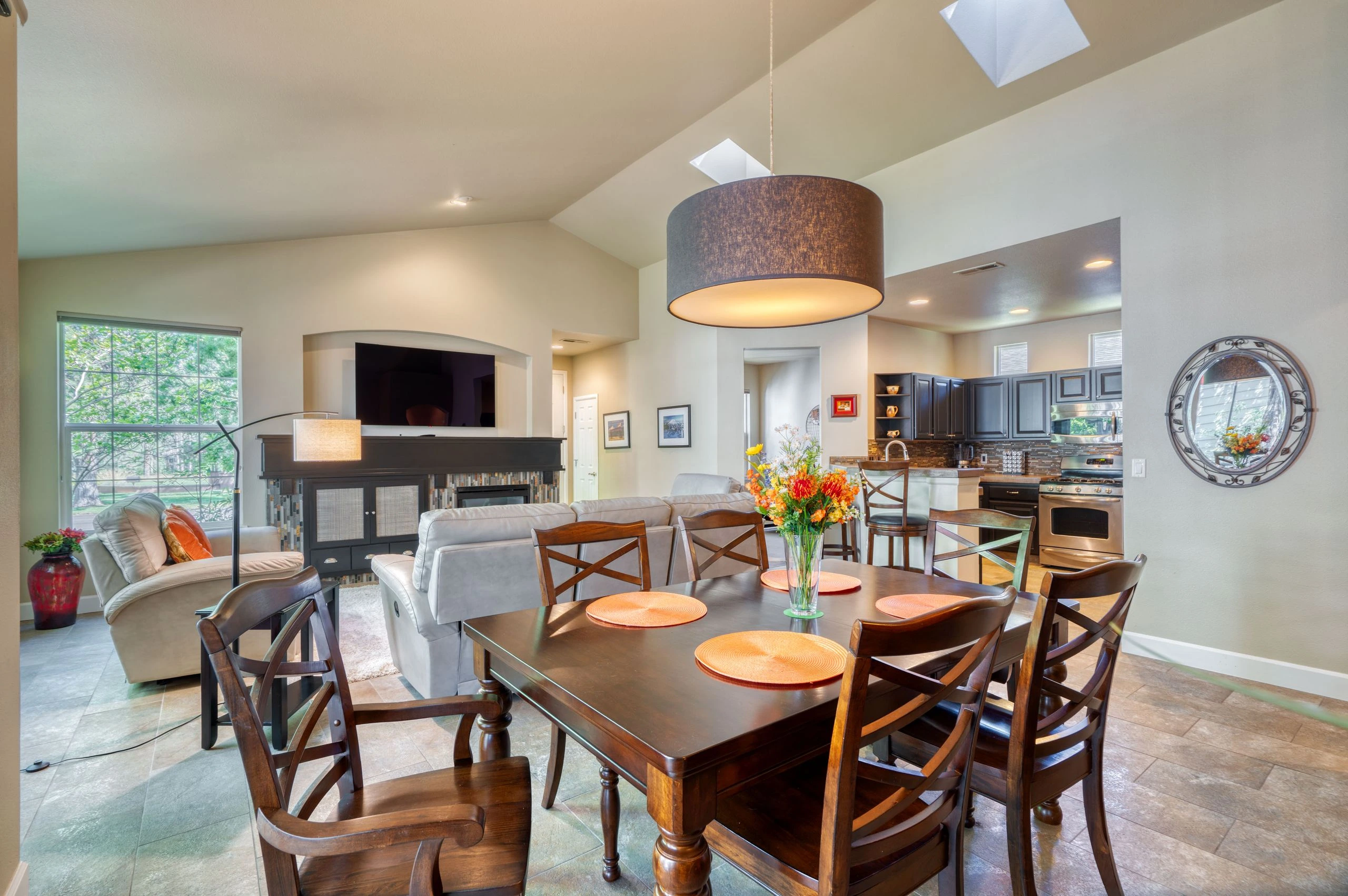 A dining table with wooden chairs sits before a living area and kitchen, with a sofa, fireplace, television, dark cabinets, and stainless steel appliances.