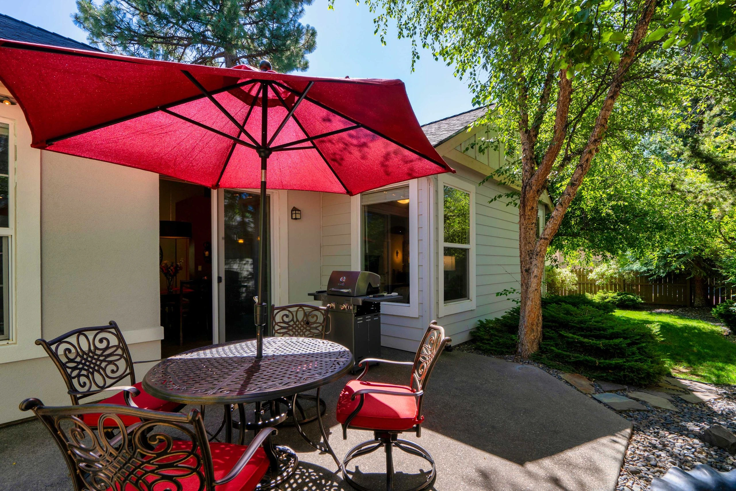 A patio with a red umbrella over a metal table and chairs, beside a house, grill, and leafy trees.