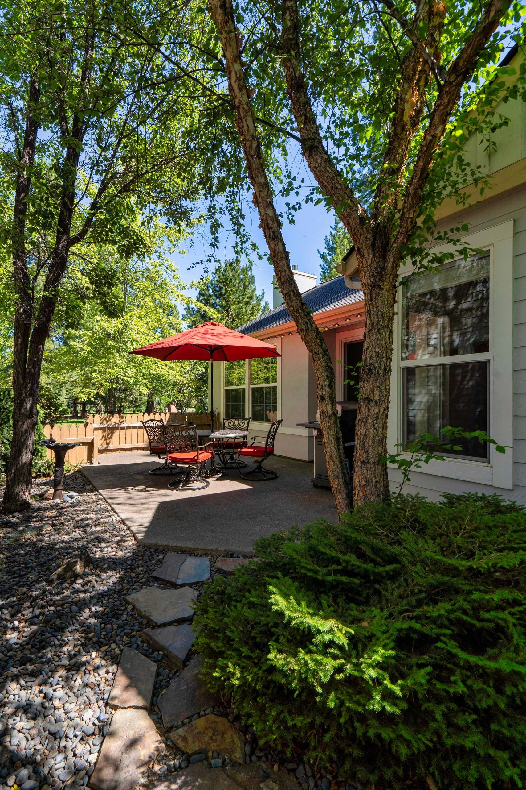 A paved patio with a red umbrella over a metal table and chairs, surrounded by trees beside a house.