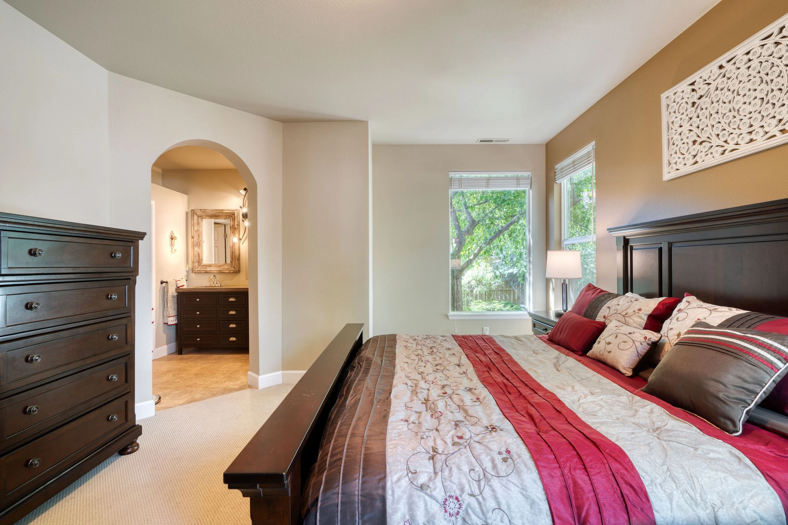 A bedroom with a large bed, dark wooden dressers, two windows, and an arched opening to a vanity area.