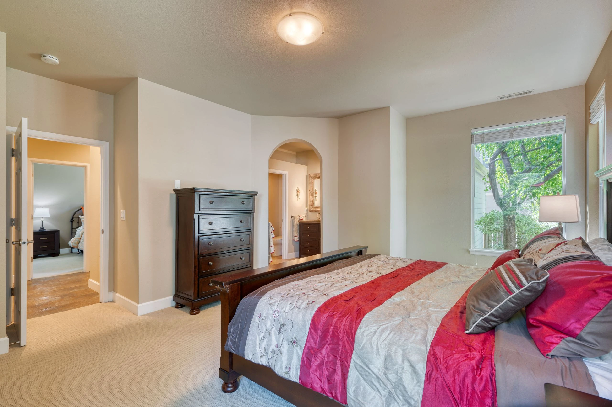 A bedroom with a large bed covered in gray and red bedding, a dark wooden dresser, and a wide window overlooking leafy trees.