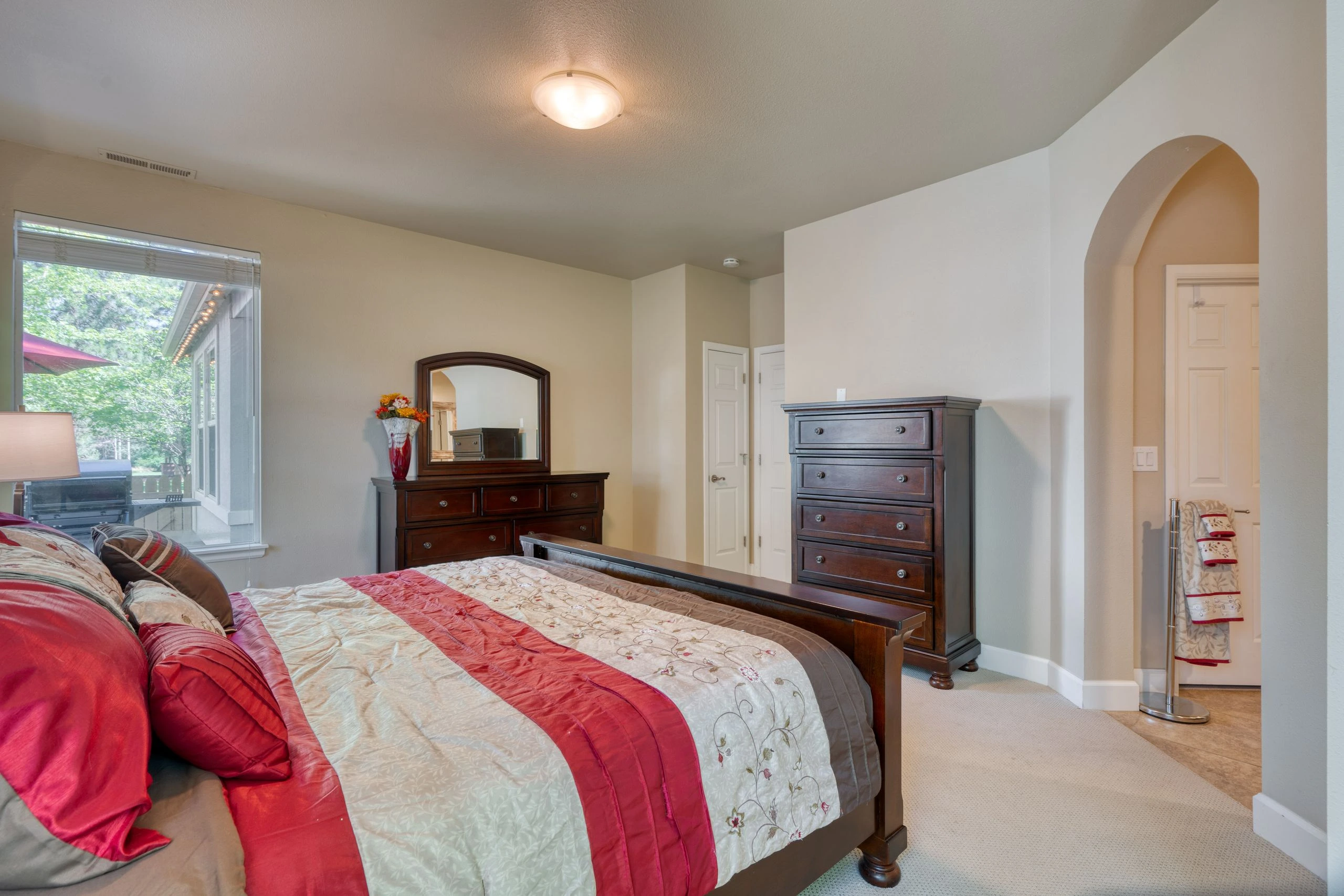 A bedroom with a bed covered in floral bedding and red accents, dark wood dressers, a wall mirror, and a large window with blinds.