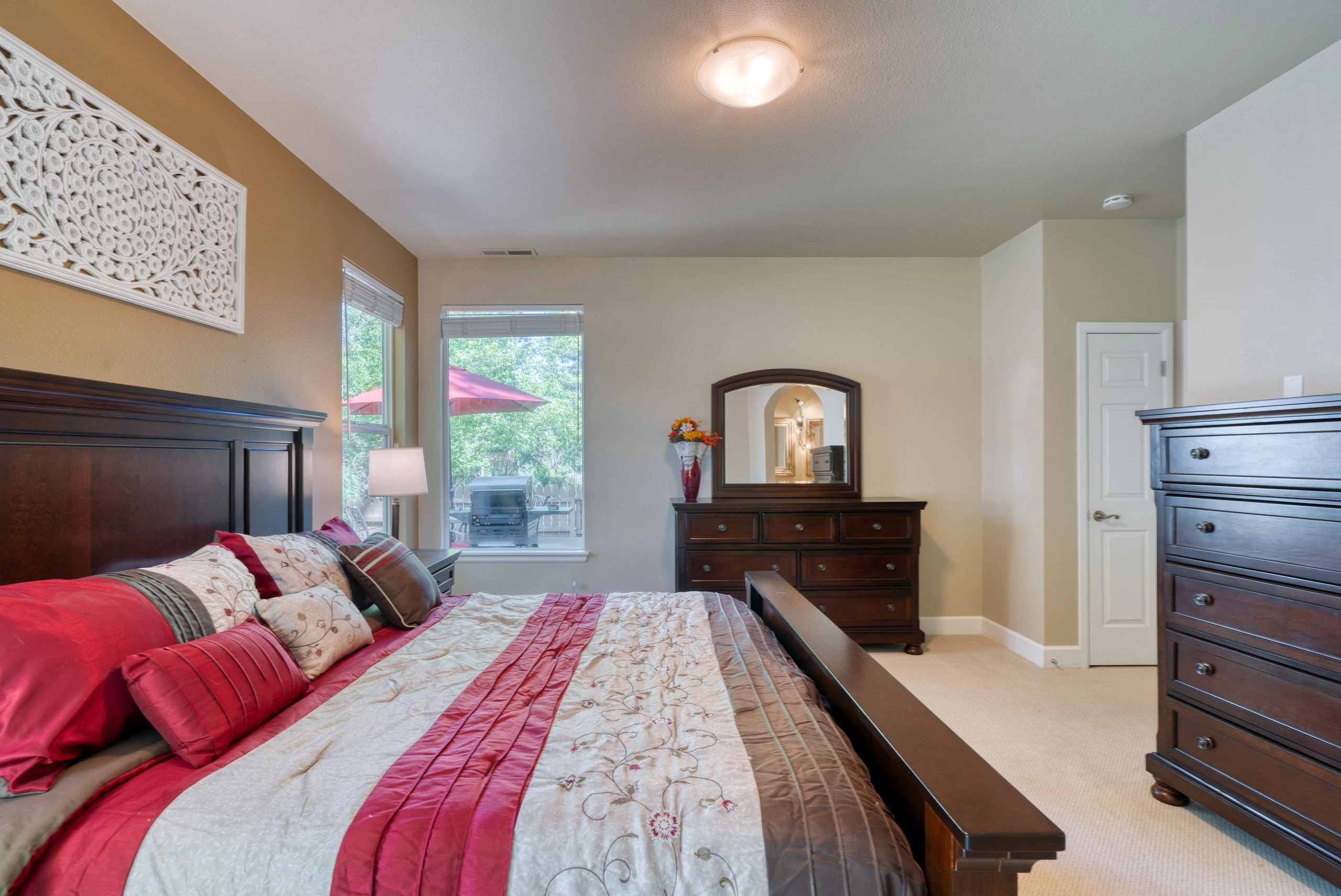 A bed with a red, cream, and brown quilt fills the foreground; a dresser and mirror stand between two windows.