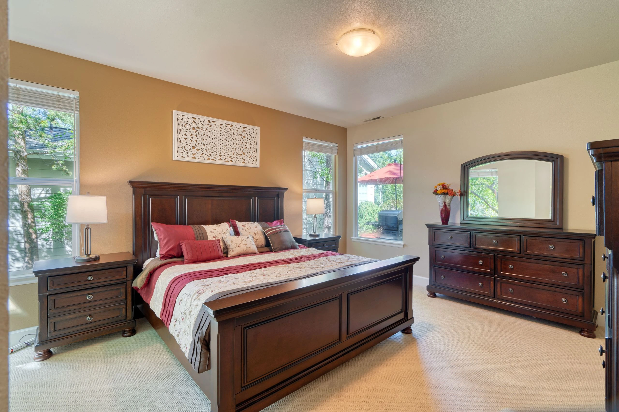 A bedroom with a dark wood bed, matching nightstand and dresser, patterned bedding, and several windows with blinds.