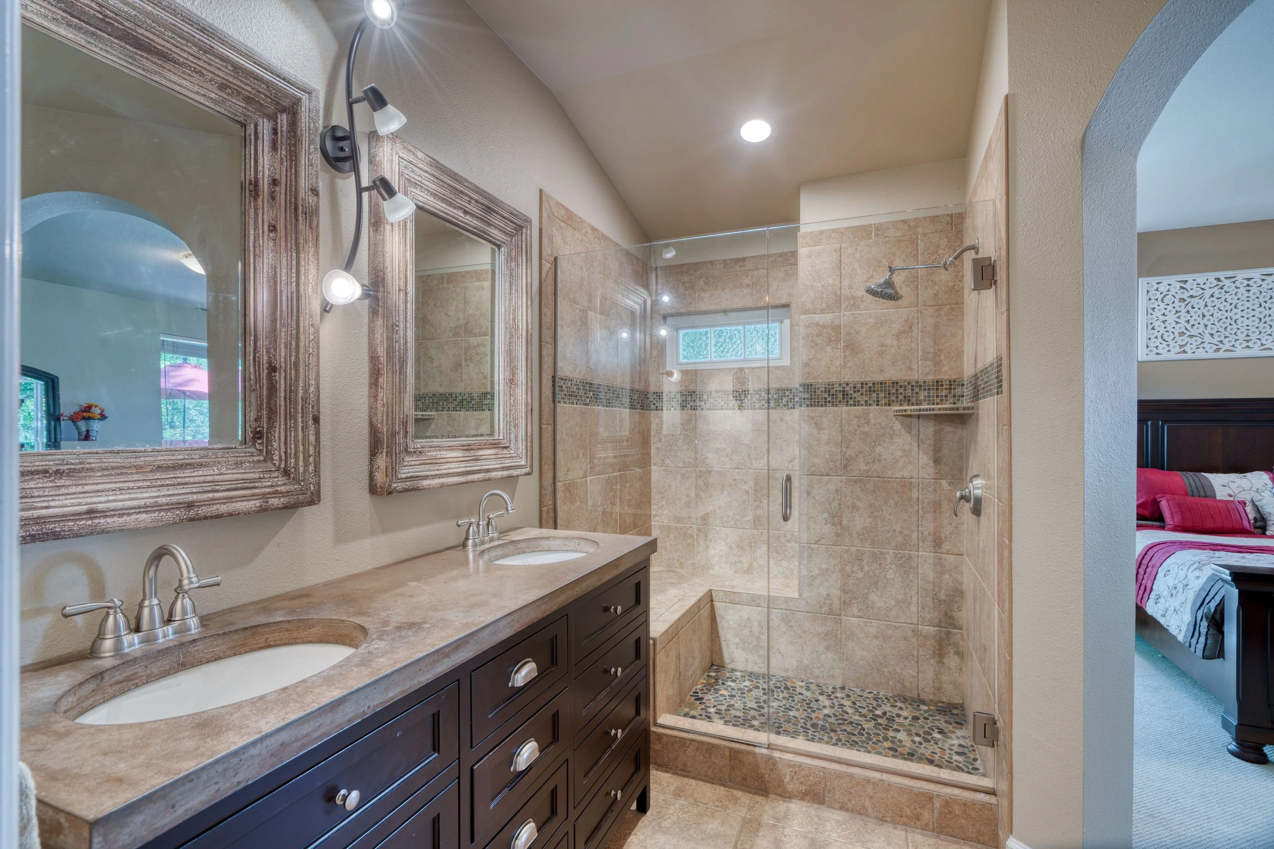 A bathroom with a dark double-sink vanity, framed mirrors, and a glass shower with tan tile and a pebble floor.