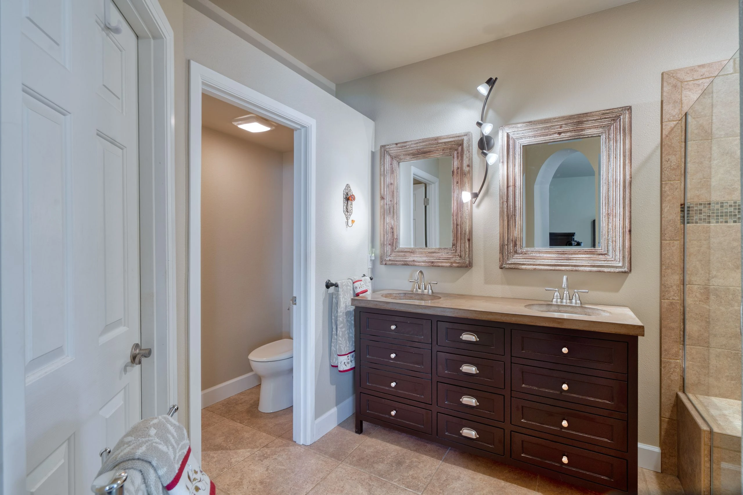 A bathroom with a dark double-sink vanity, two framed mirrors, and a tiled shower beside an open doorway to a toilet.
