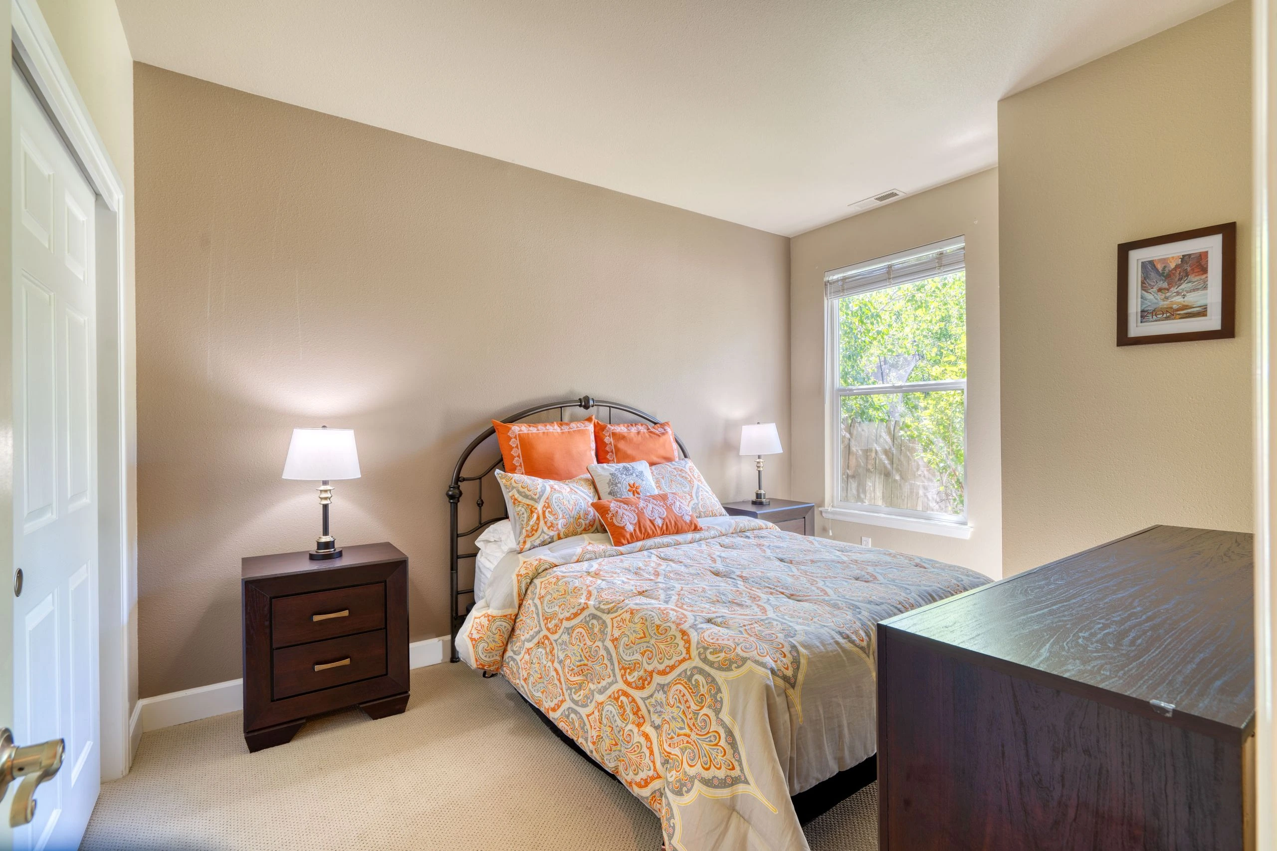 A bedroom with a patterned bed, two bedside tables and lamps, a window with blinds, and framed wall art.