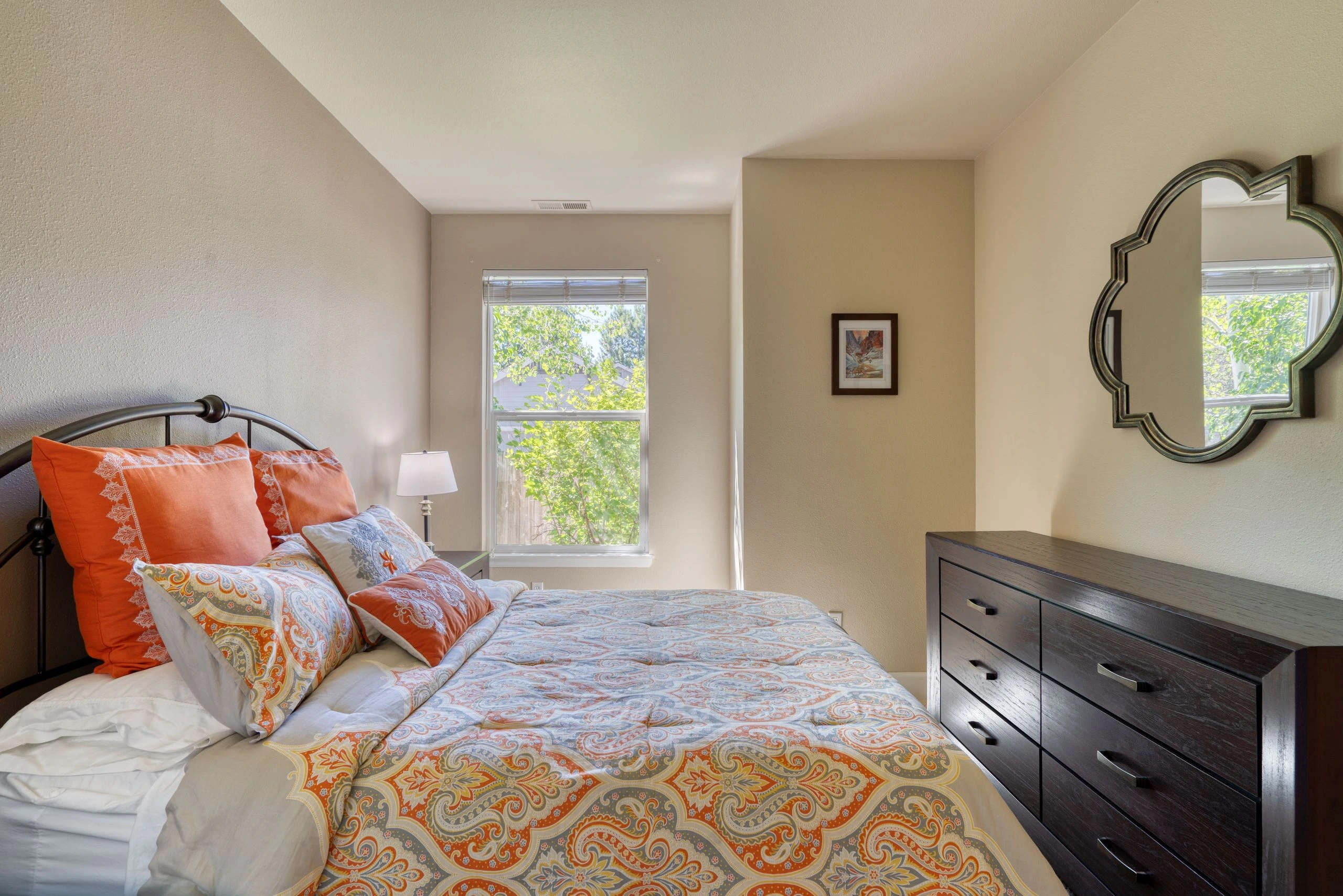A bed with patterned bedding and orange pillows sits beneath a window, beside a dark dresser and decorative mirror.