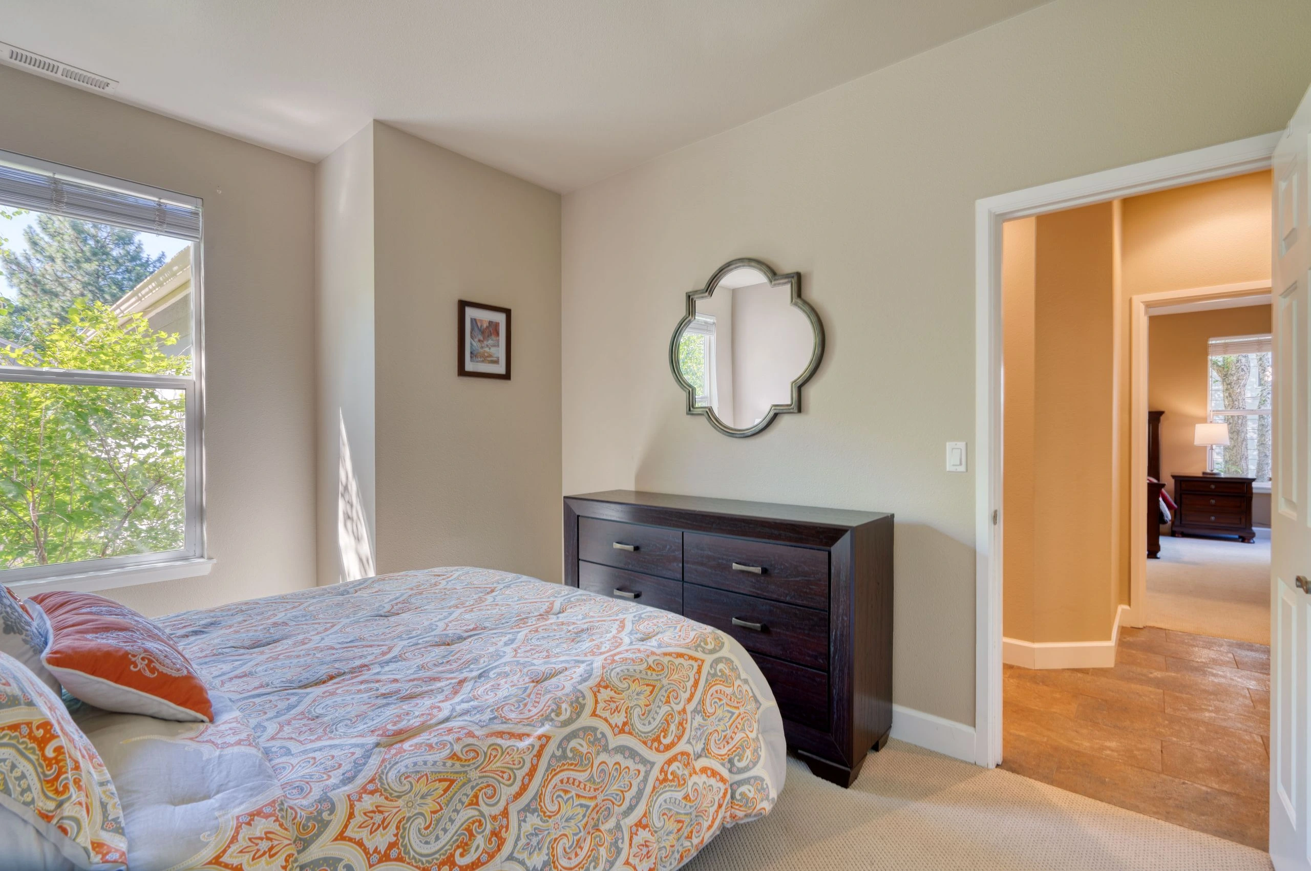 A bedroom with a patterned bed, dark dresser, decorative wall mirror, and window with blinds.