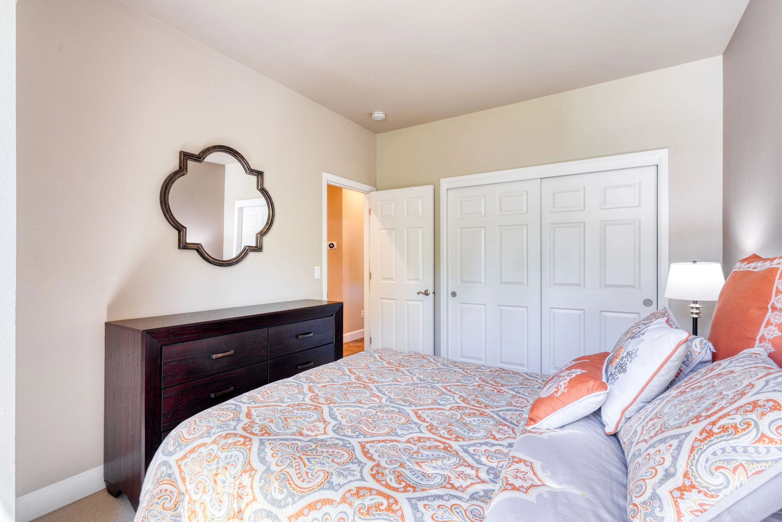 A bed with patterned bedding and pillows sits beside a dark dresser and ornate mirror; white doors and a lit lamp are visible.