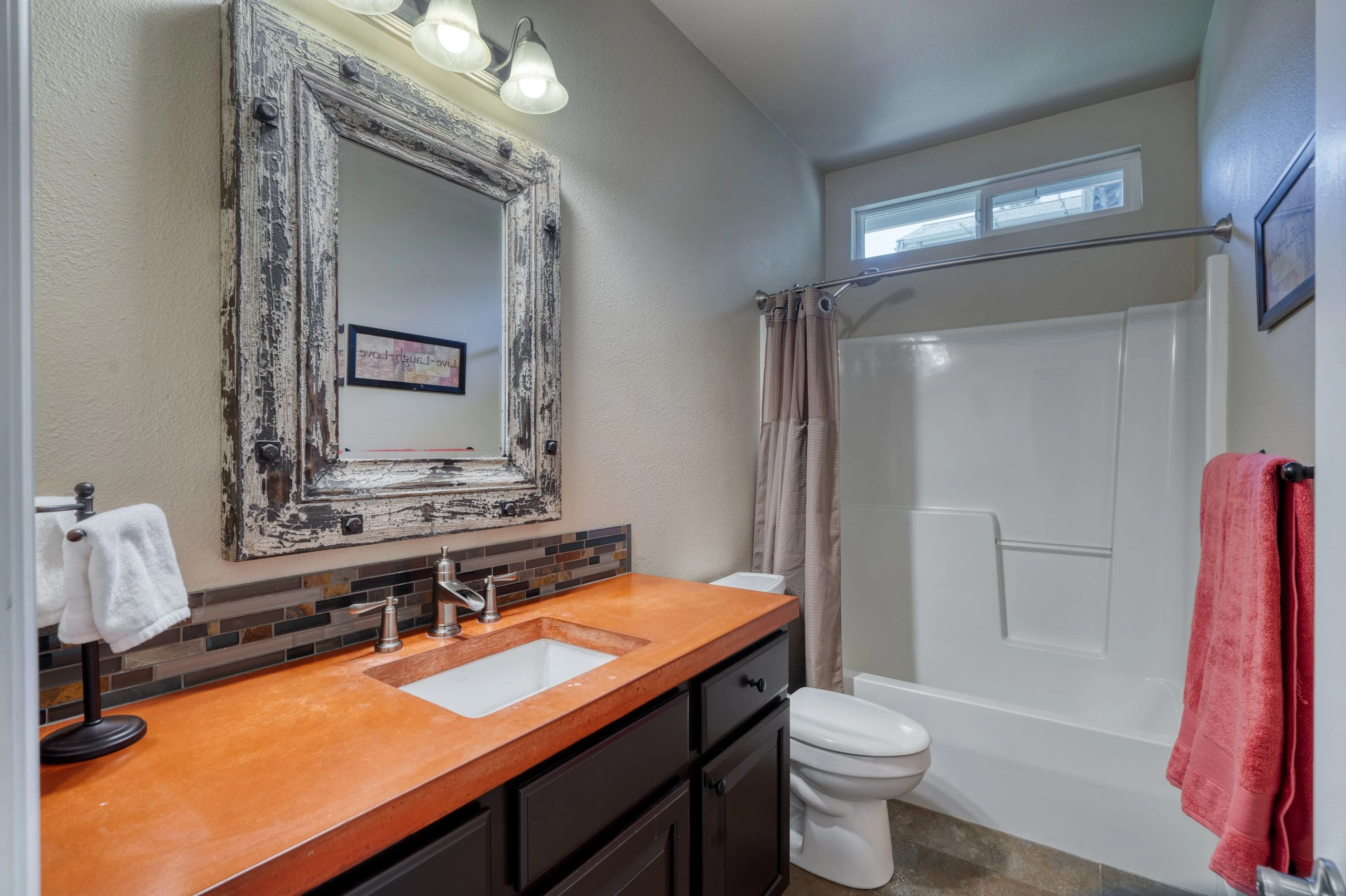 Bathroom with an orange counter and white sink, distressed-framed mirror, dark cabinets, toilet, and bathtub with a curtain.