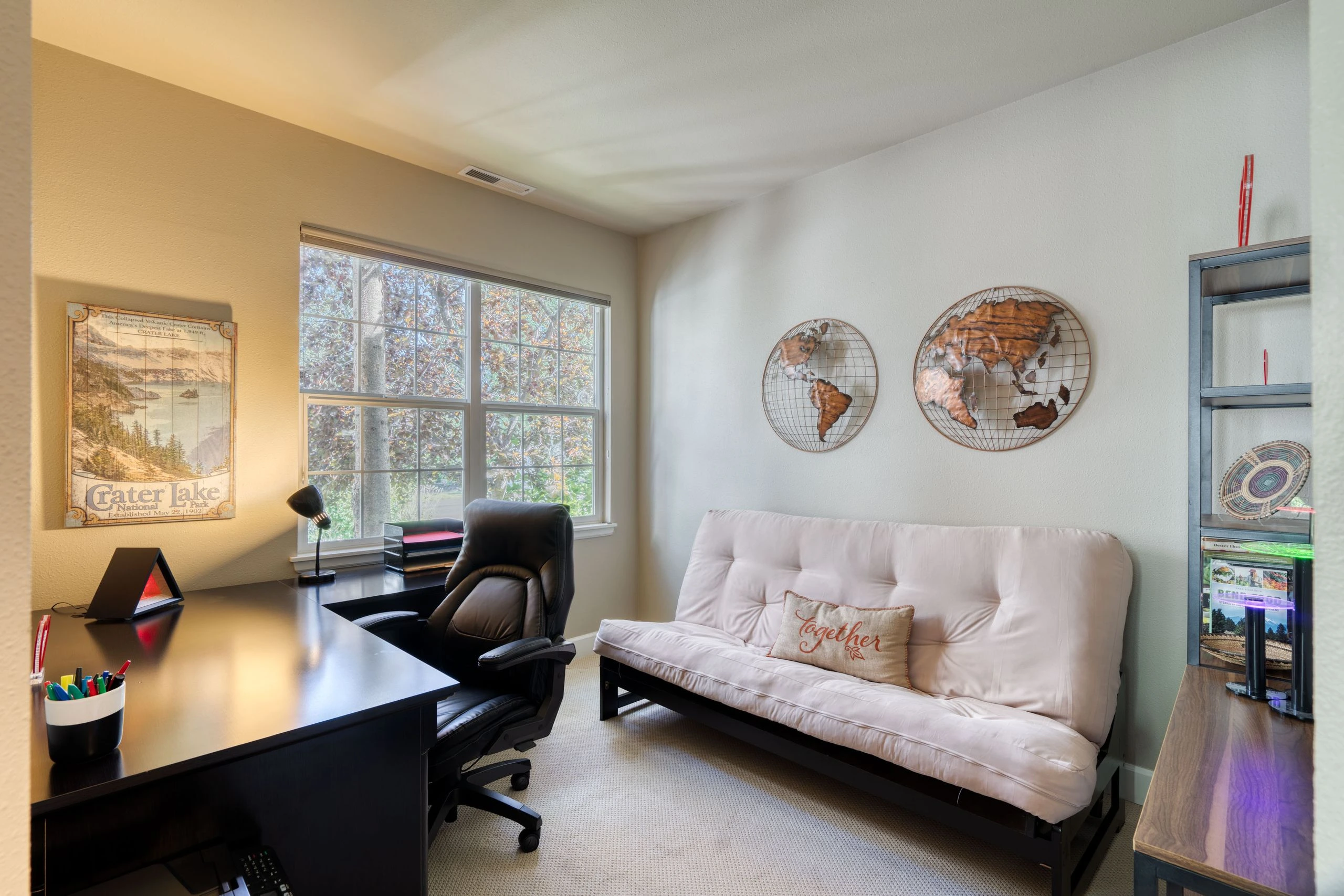A room with a black desk and office chair beside a pale futon, large windows, and two metal world map wall decorations.