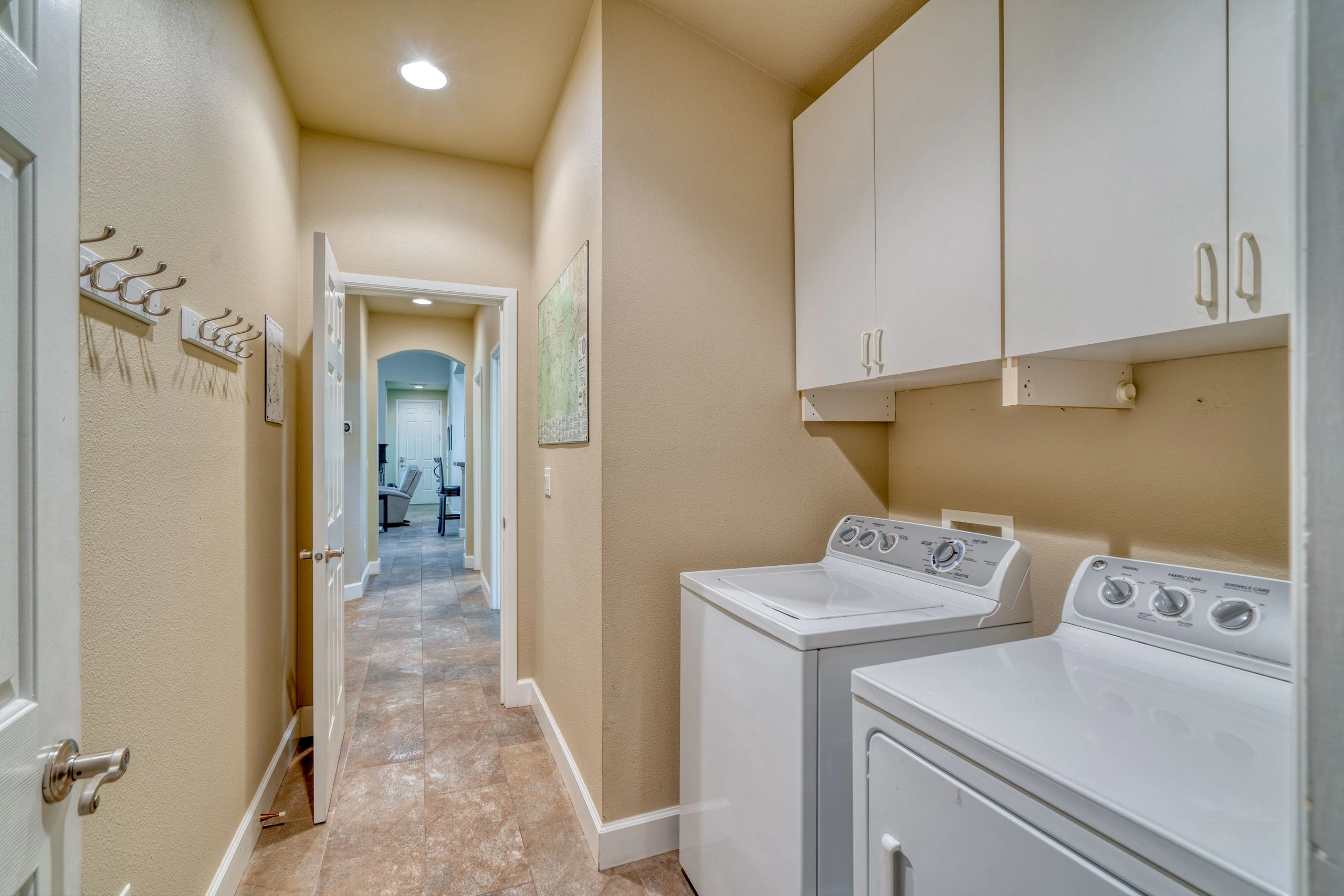 A narrow laundry room has two white appliances, overhead cabinets, wall hooks, and a tiled floor leading to an open doorway.