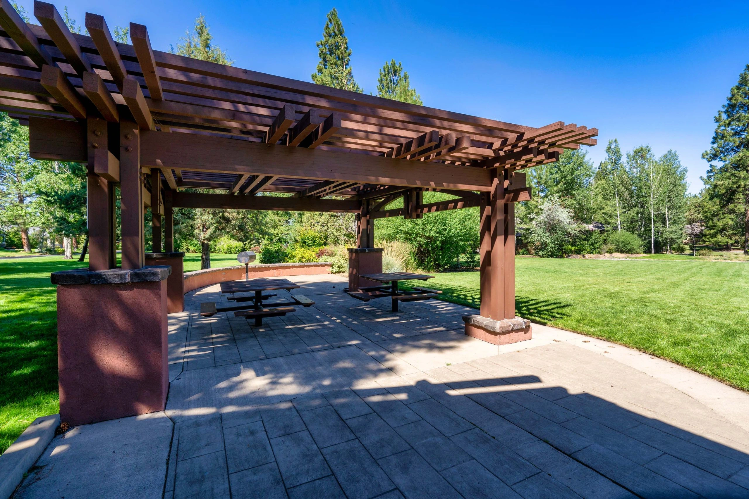 A large wooden pergola shelters picnic tables on a paved patio surrounded by lawn and trees.