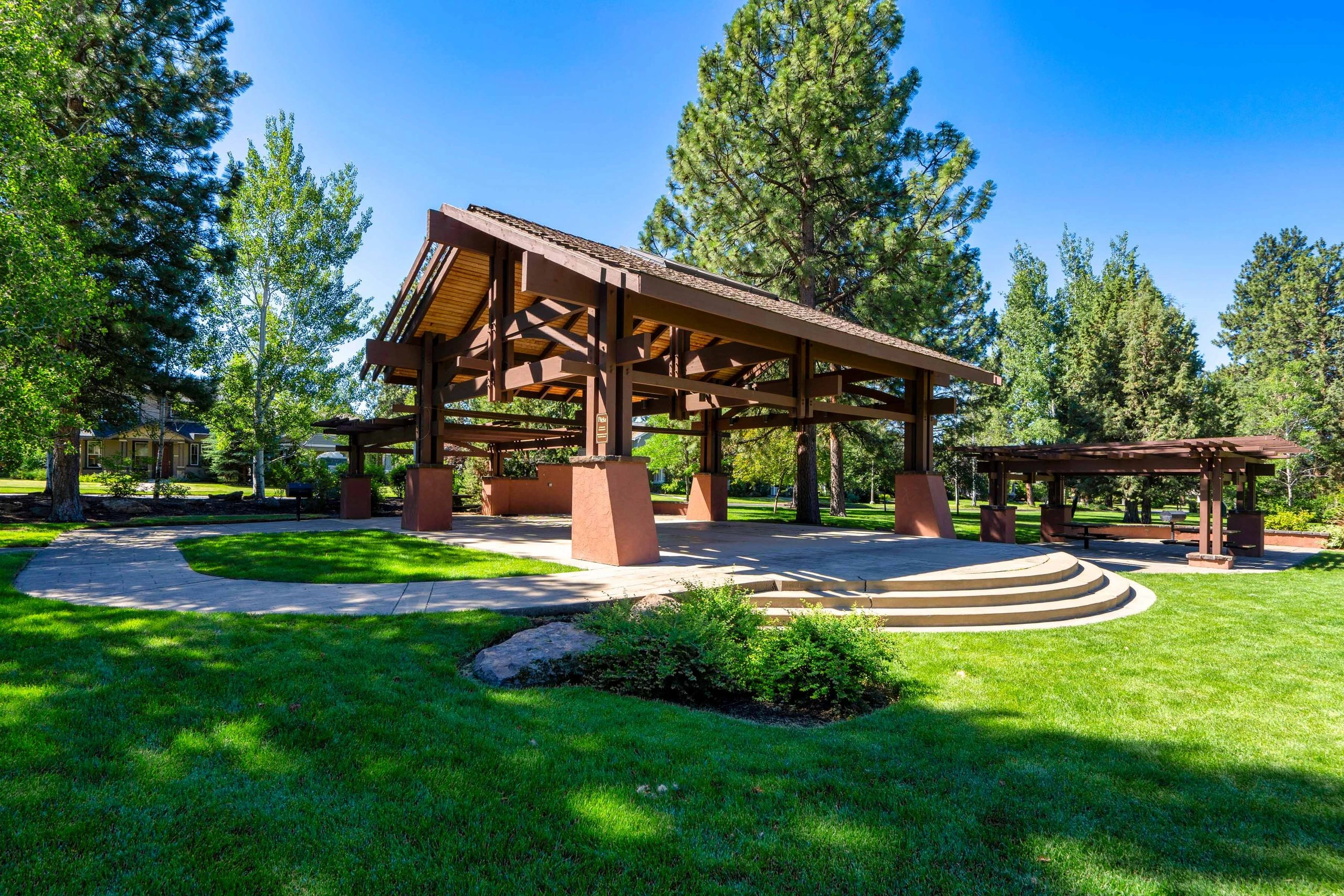 A timber pavilion with a broad sloping roof stands among trees beside paved paths and curved steps.