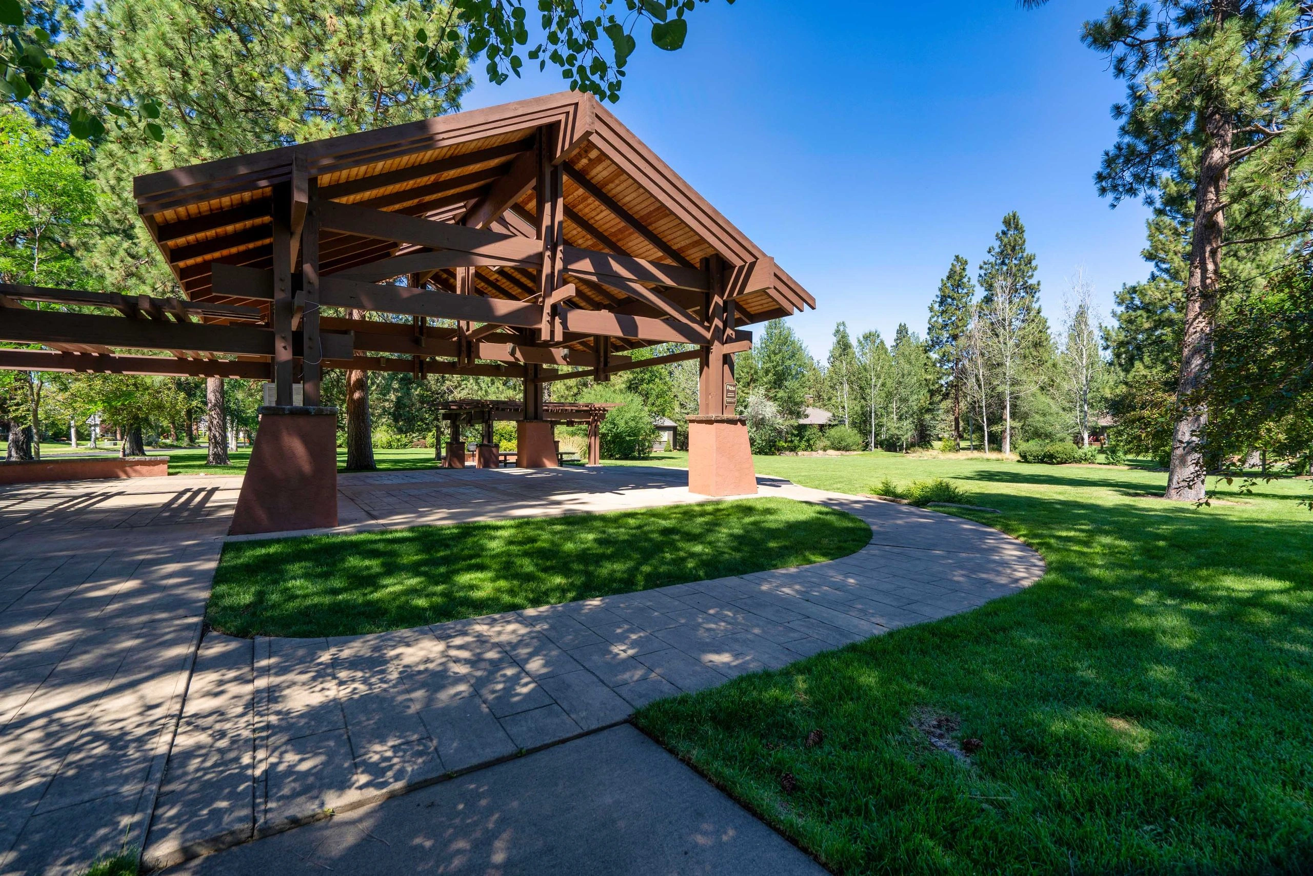 A large timber pavilion with a peaked roof stands beside paved paths and lawns among trees.