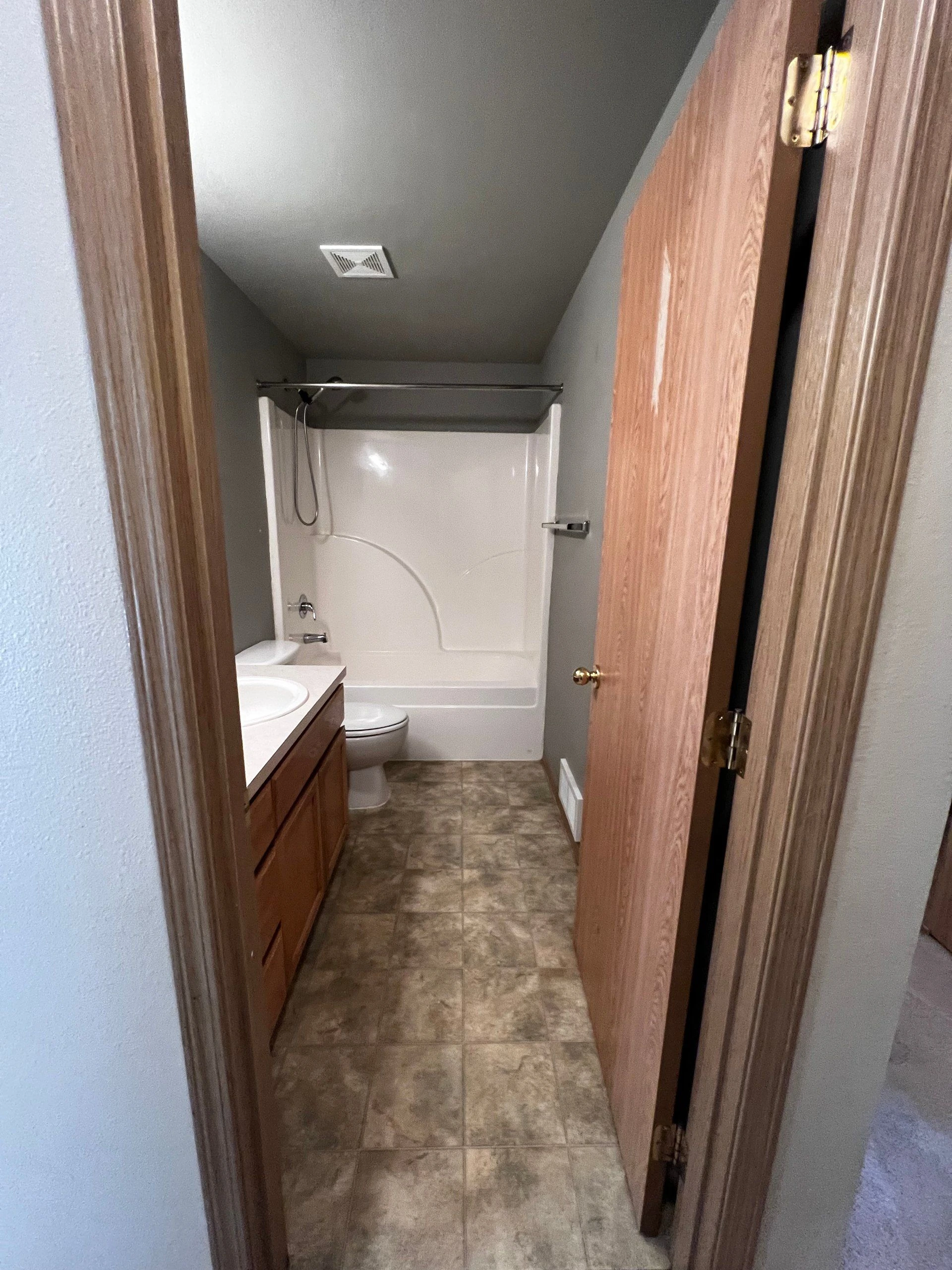 A narrow bathroom with a white tub and shower, toilet, wood vanity with sink, and patterned tile floor.