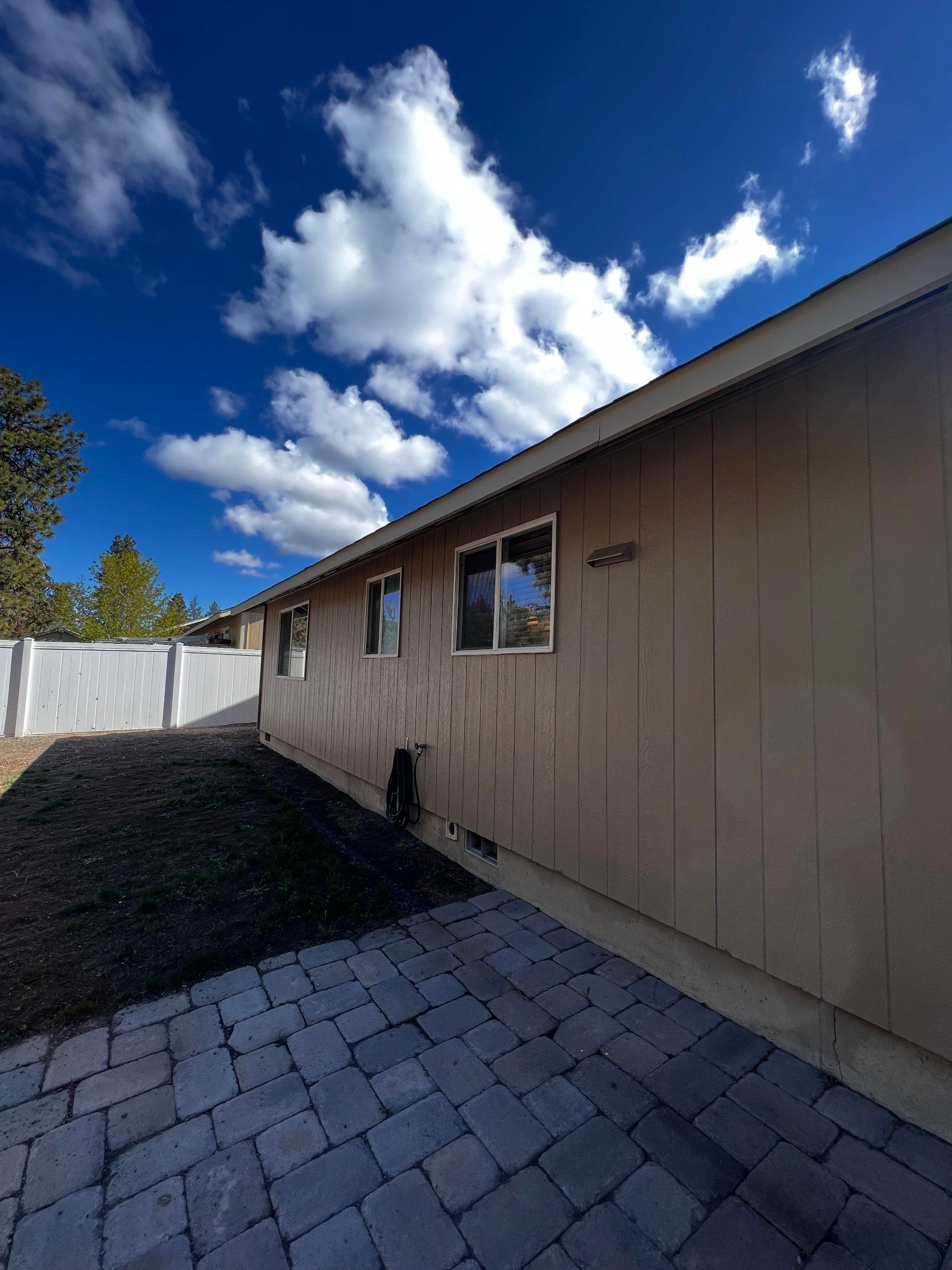 A brown-sided building with windows stands beside a gray paved path and white fence under a blue, cloudy sky.