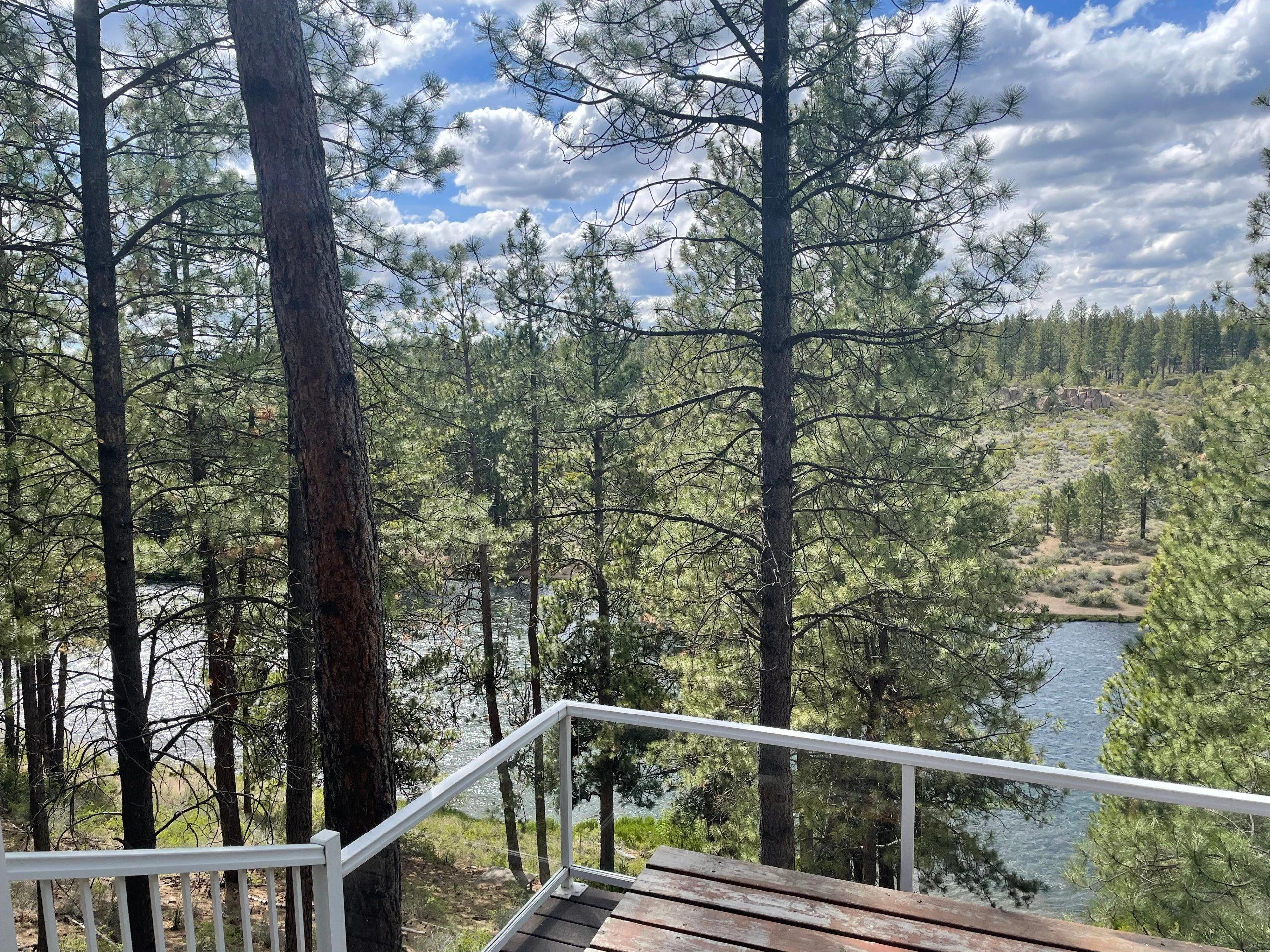 A wooden deck with white railings overlooks a river winding through pine trees beneath a partly cloudy sky.