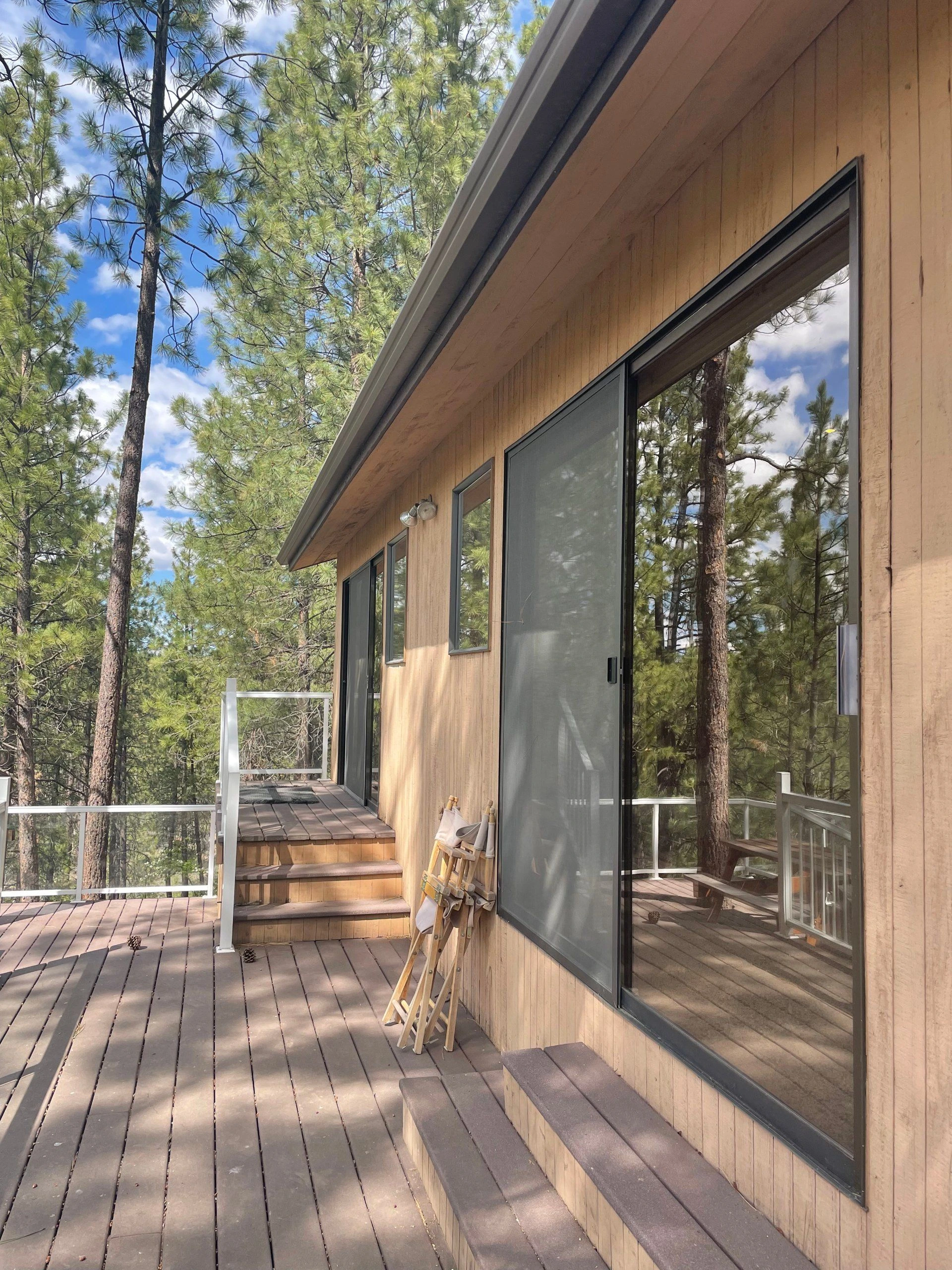 A wooden deck with steps runs alongside a wood-sided building with large glass doors, surrounded by tall trees.