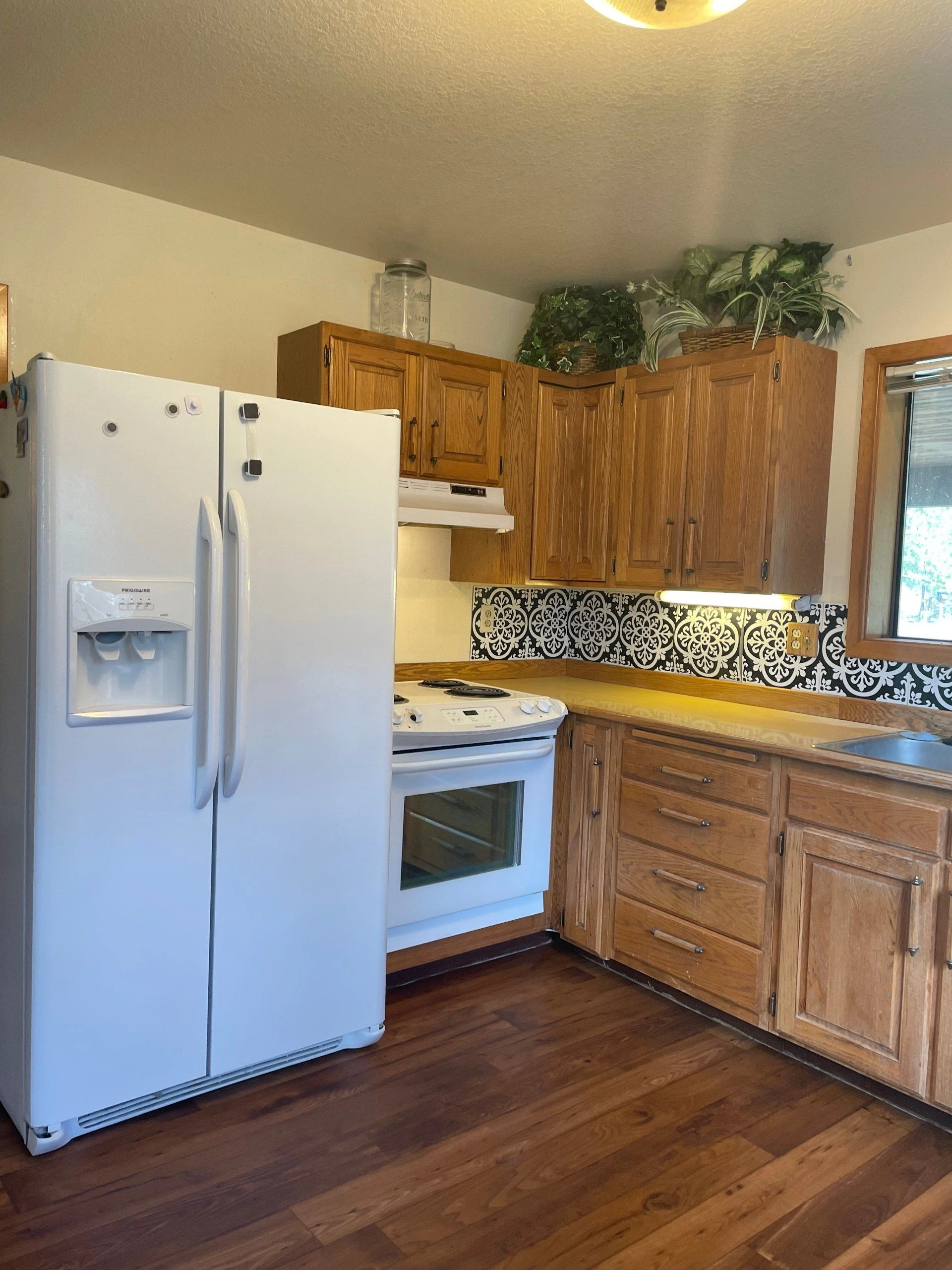 A kitchen with a white refrigerator and stove, wooden cabinets, a patterned backsplash, and potted plants above the cabinets.