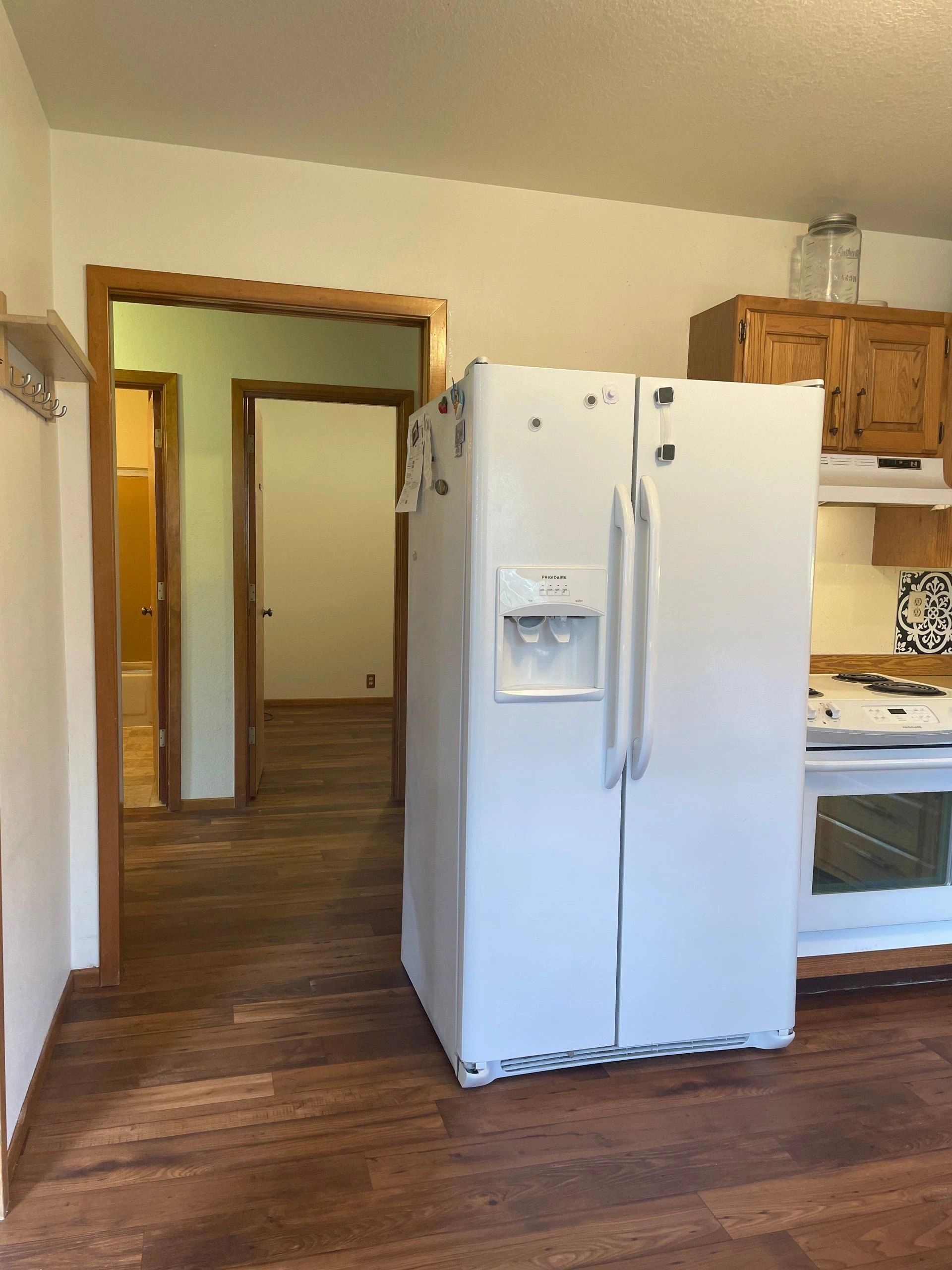 A white side-by-side refrigerator with a water dispenser stands beside a white stove and wooden cabinets.