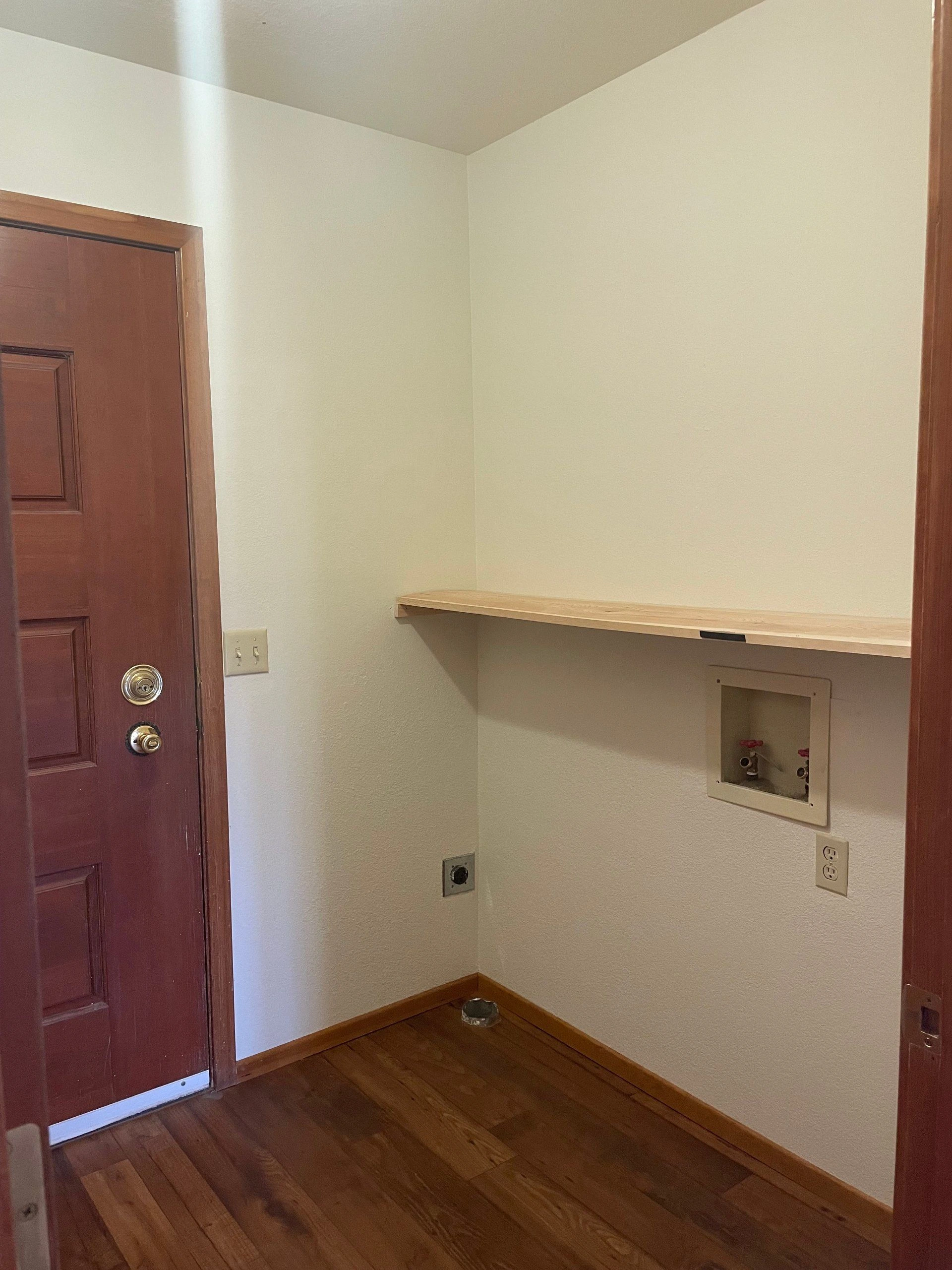 A small room with a dark wooden door, a long wall shelf, and a recessed utility box above the wood floor.
