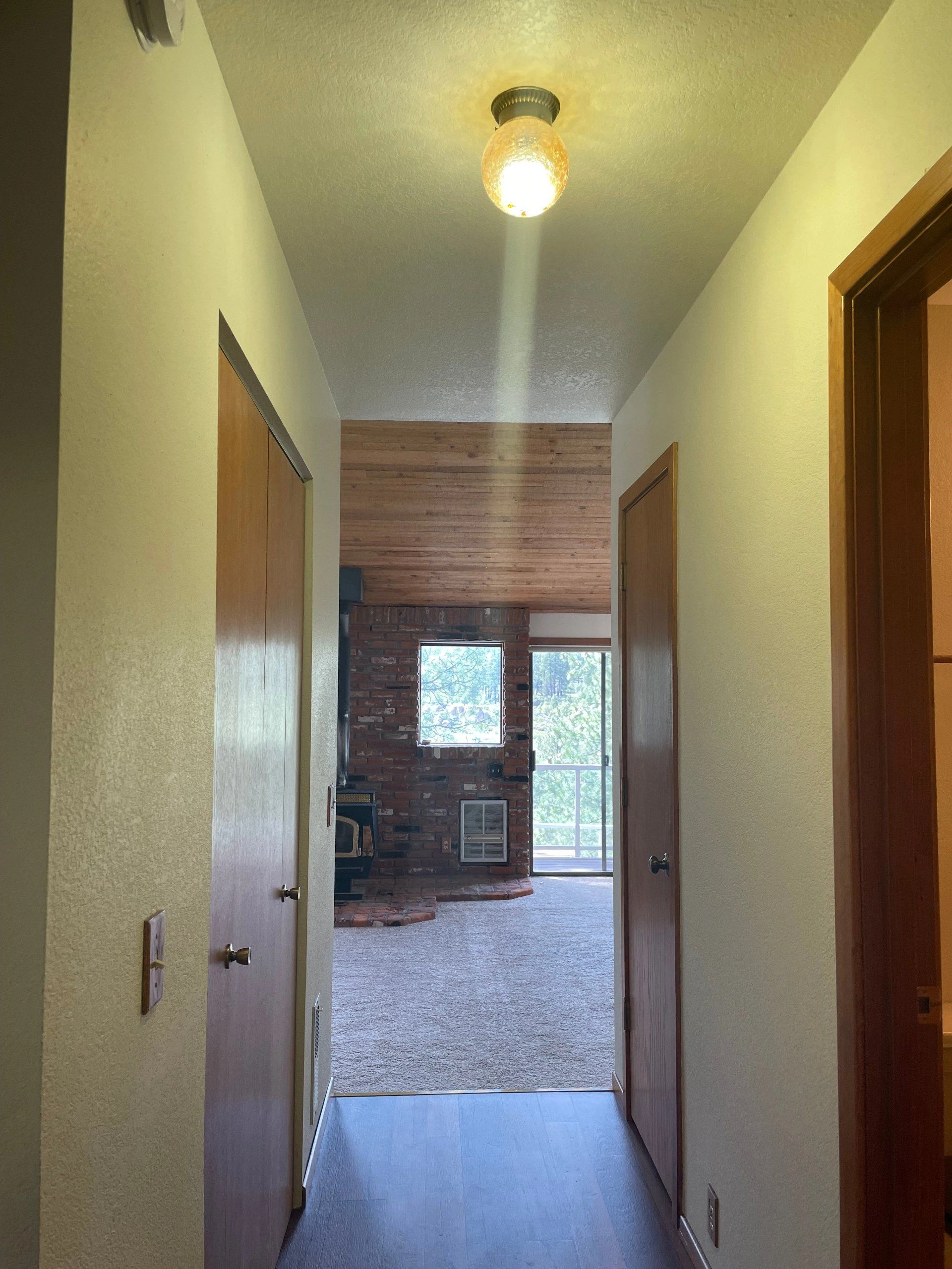 A narrow hallway with open wooden doors leads to a carpeted room with a brick wall, wood-paneled ceiling, and windows.