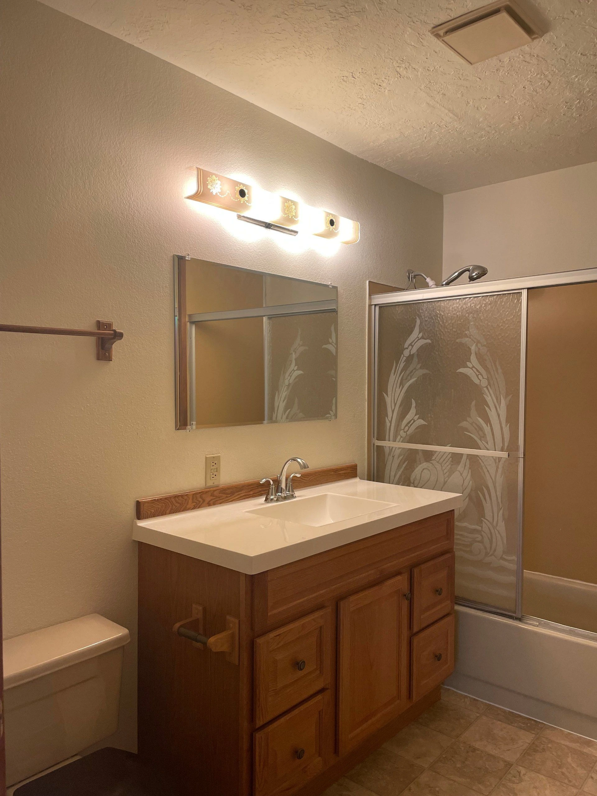 A bathroom with a wood vanity and white sink, wall mirror, illuminated fixture, toilet, and patterned sliding shower doors.