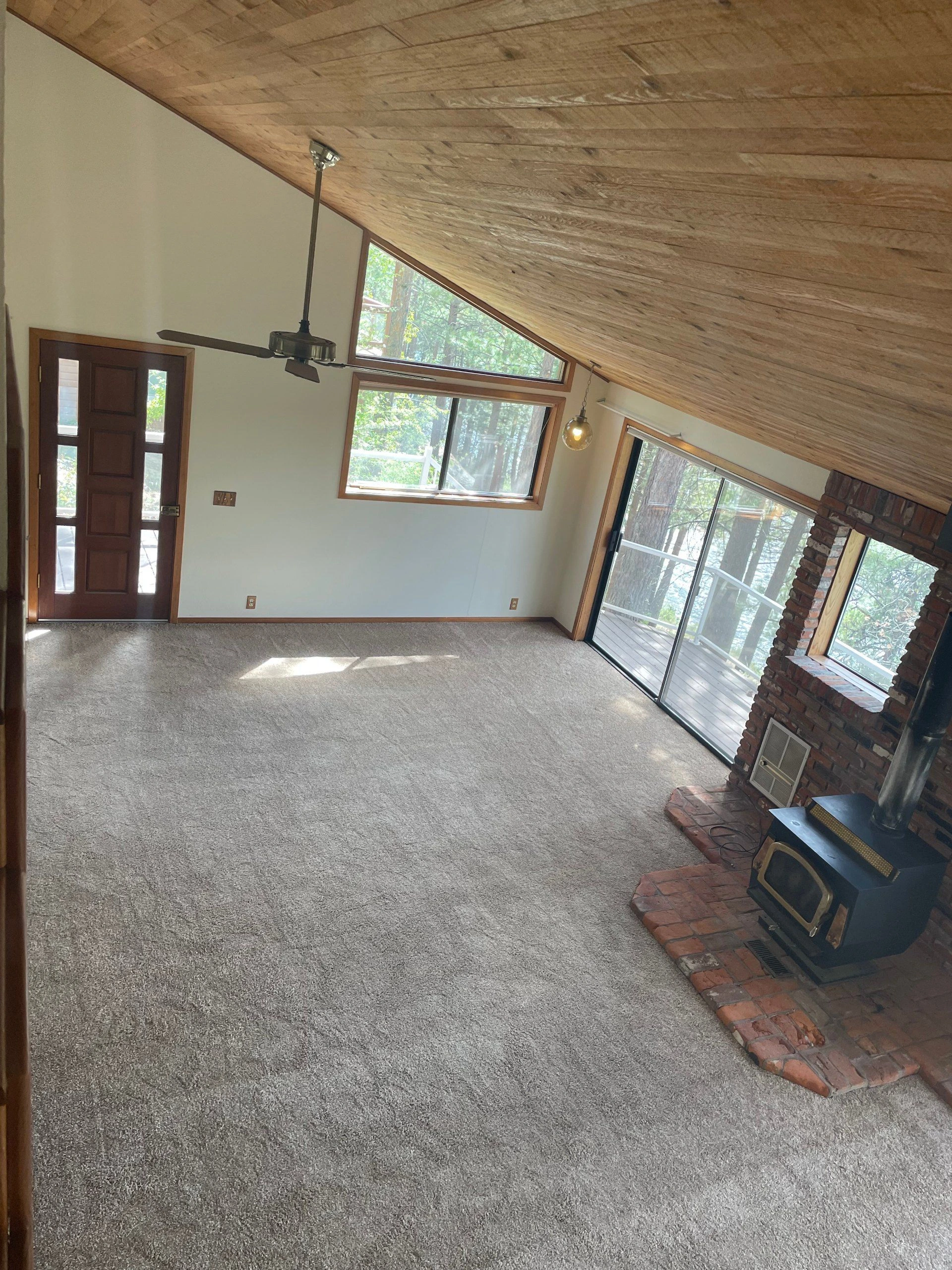 A carpeted room has a sloped wood-plank ceiling, ceiling fan, large windows, a dark wood door, and a brick fireplace with a stove.