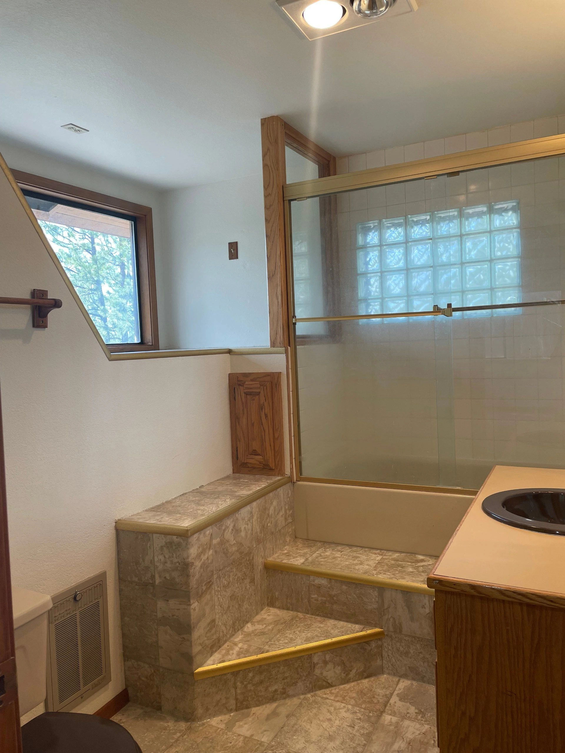 A tiled bathing area has a glass shower enclosure, built-in steps, a wood-framed window, and part of a sink counter.