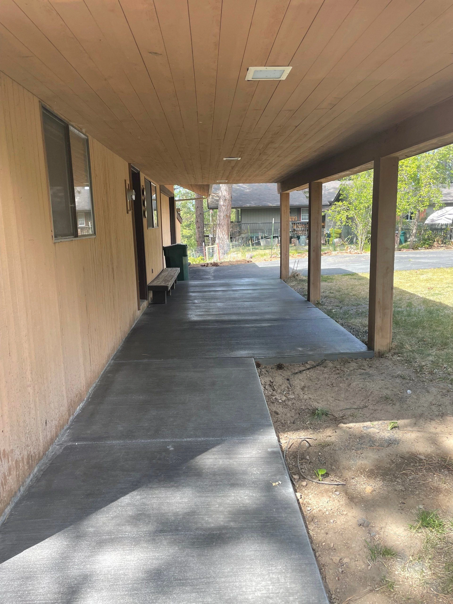 A covered concrete walkway runs beside a tan-sided building, with wooden support posts along the open side.