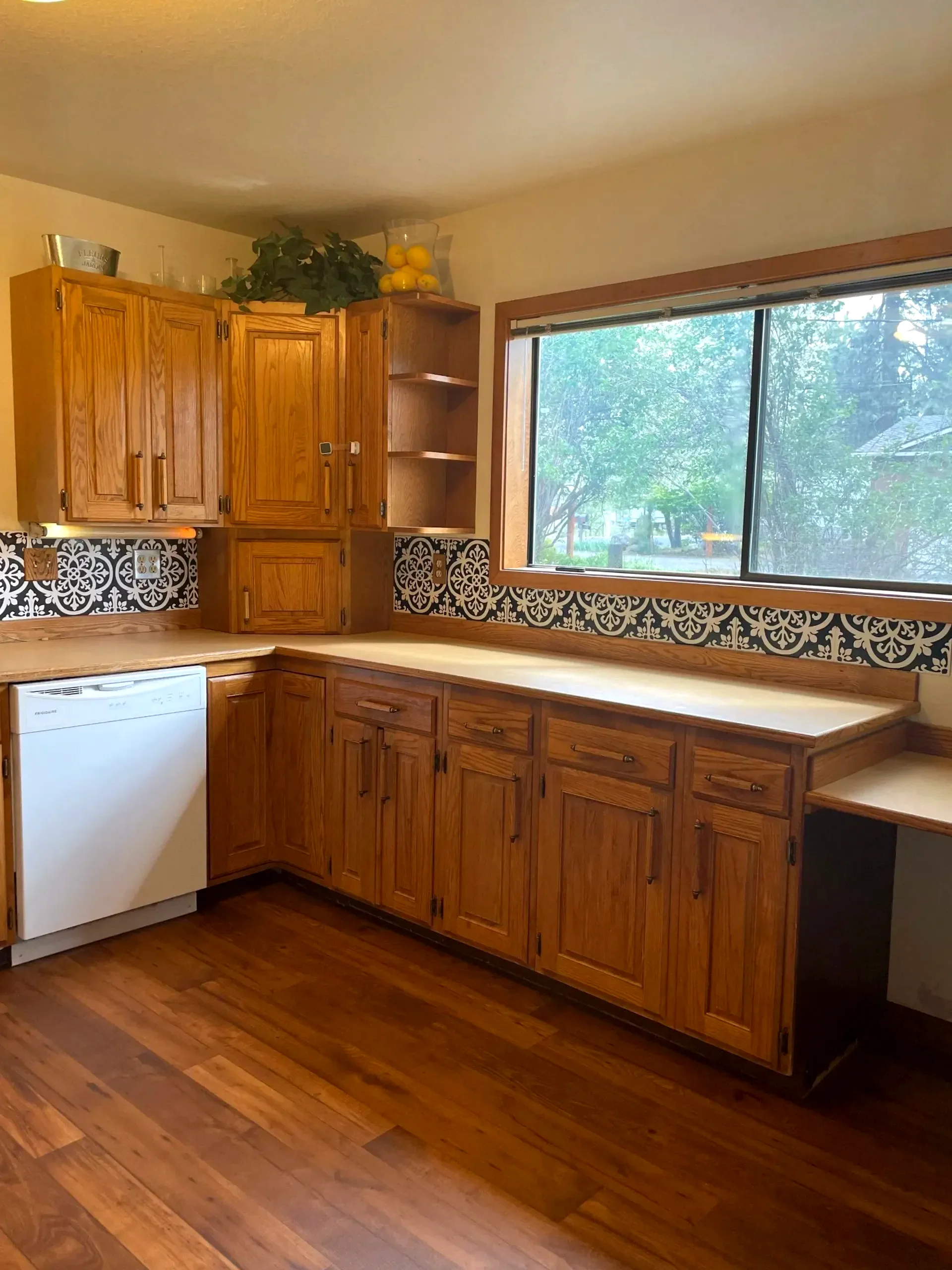 A kitchen with wood cabinets, a patterned backsplash, a large window, and a white dishwasher.