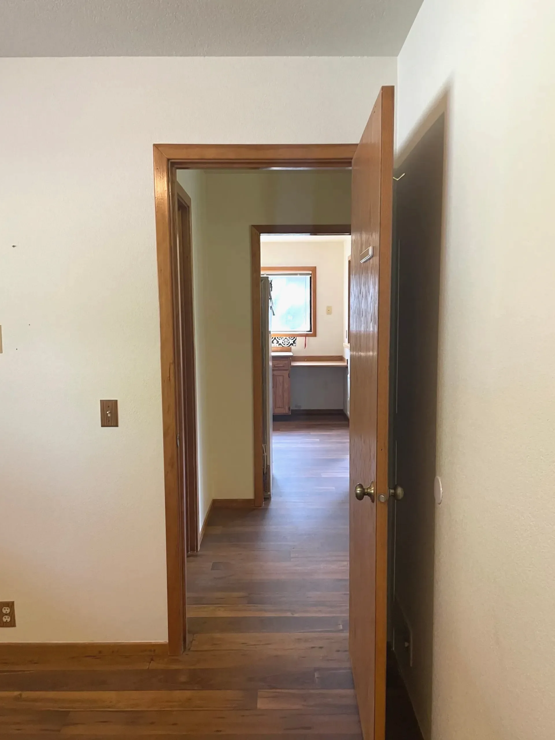 An open wooden door frames a hallway with wood flooring and a window at the far end.