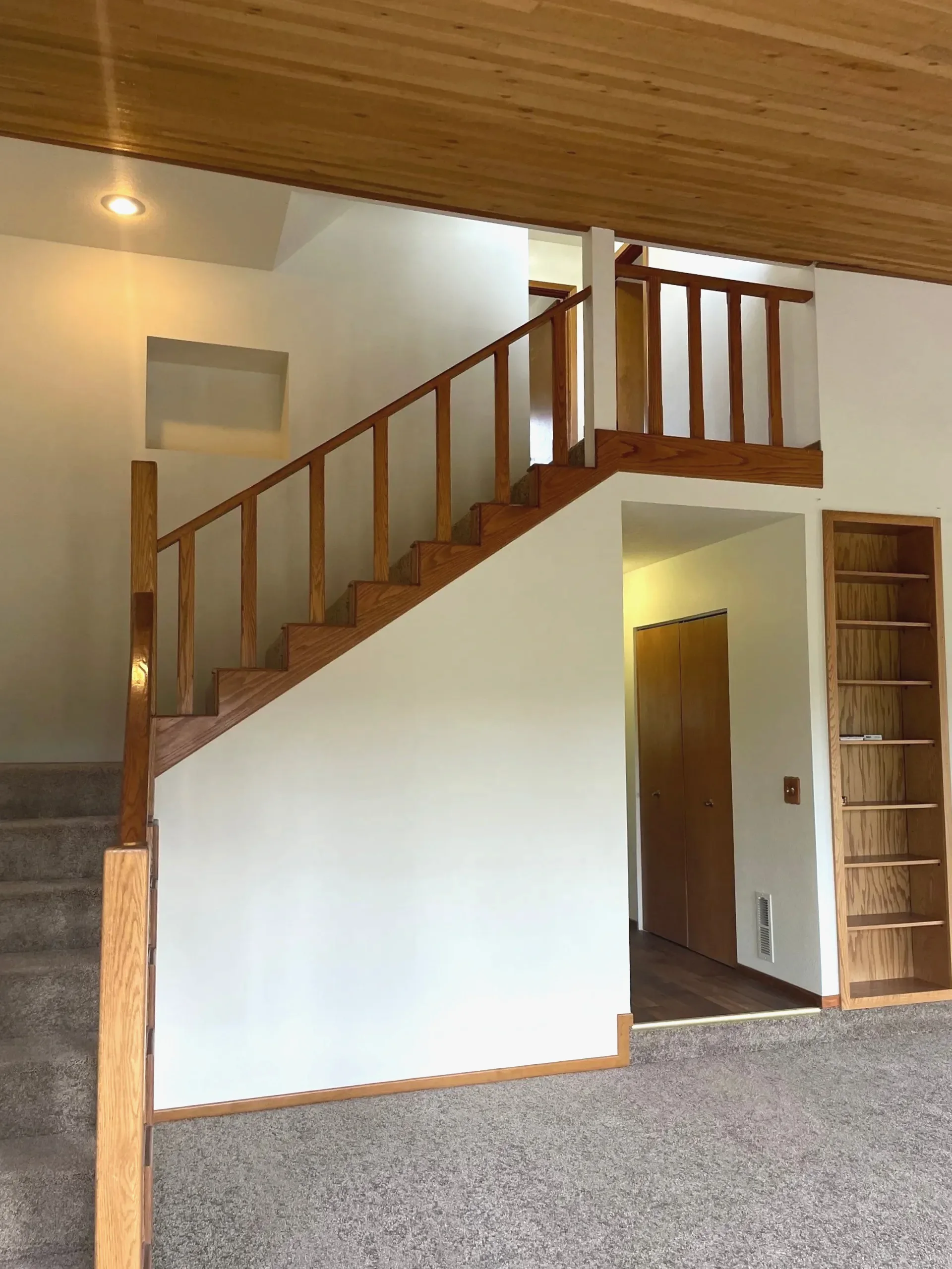 A carpeted staircase with wooden railings rises beneath a wood-paneled ceiling; a doorway and built-in shelves are visible beside it.
