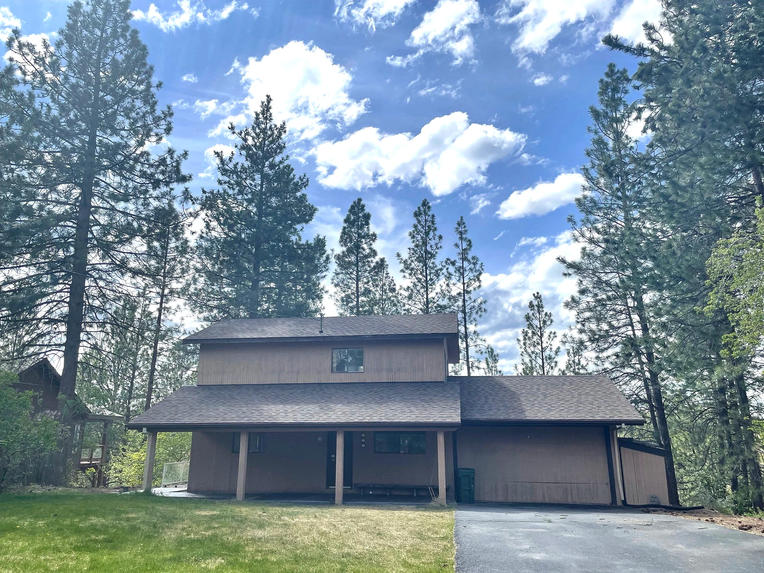 A two-story brown building with broad shingled roofs sits among tall pine trees beneath a blue, cloud-filled sky.