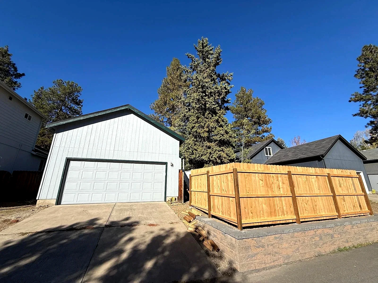 A light-colored detached garage with a double door stands beside a tall wooden fence and concrete driveway.