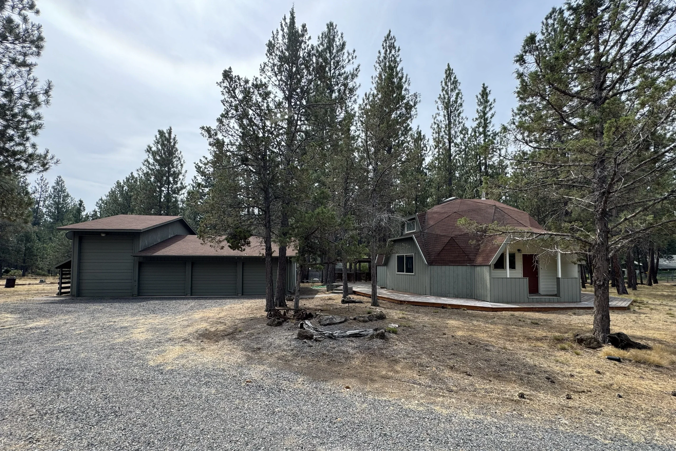 A gray house with a red-brown roof and adjoining three-door garage stands among tall pine trees beside a gravel driveway.