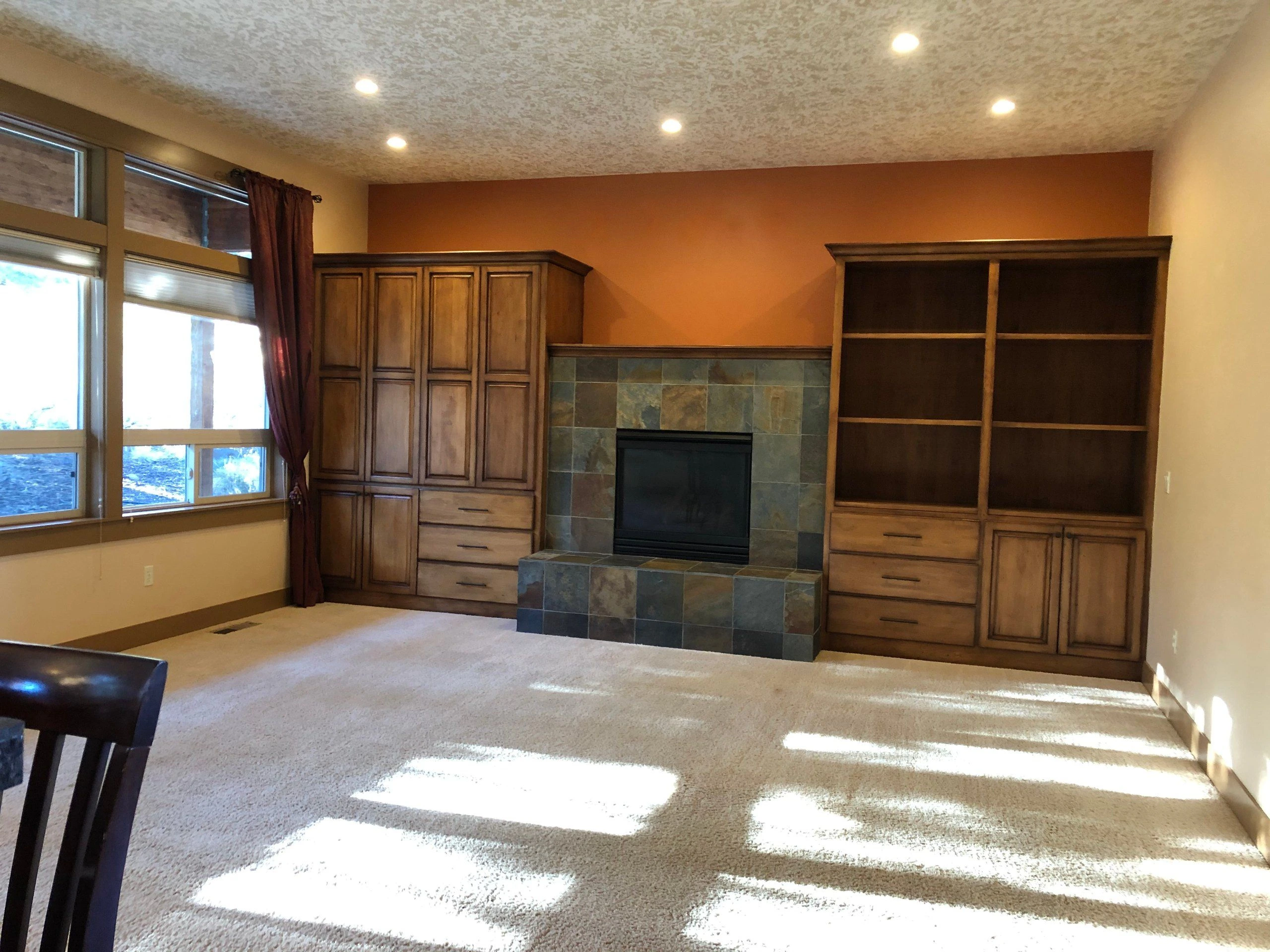 A carpeted room has a tiled fireplace between wooden cabinets and open shelves, with large windows along one wall.