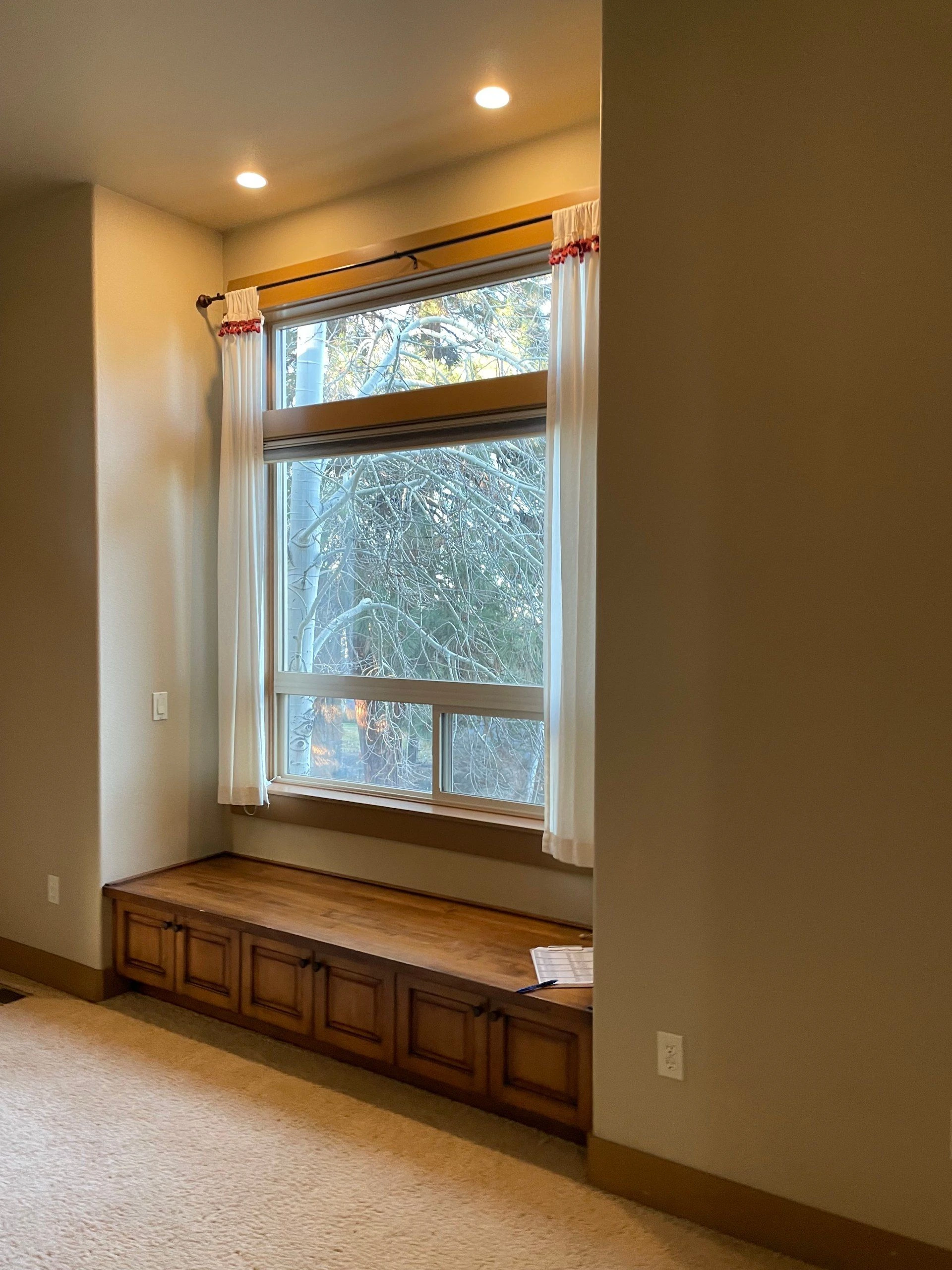A tall window with white curtains sits above a long wooden cabinet; tree branches are visible outside.
