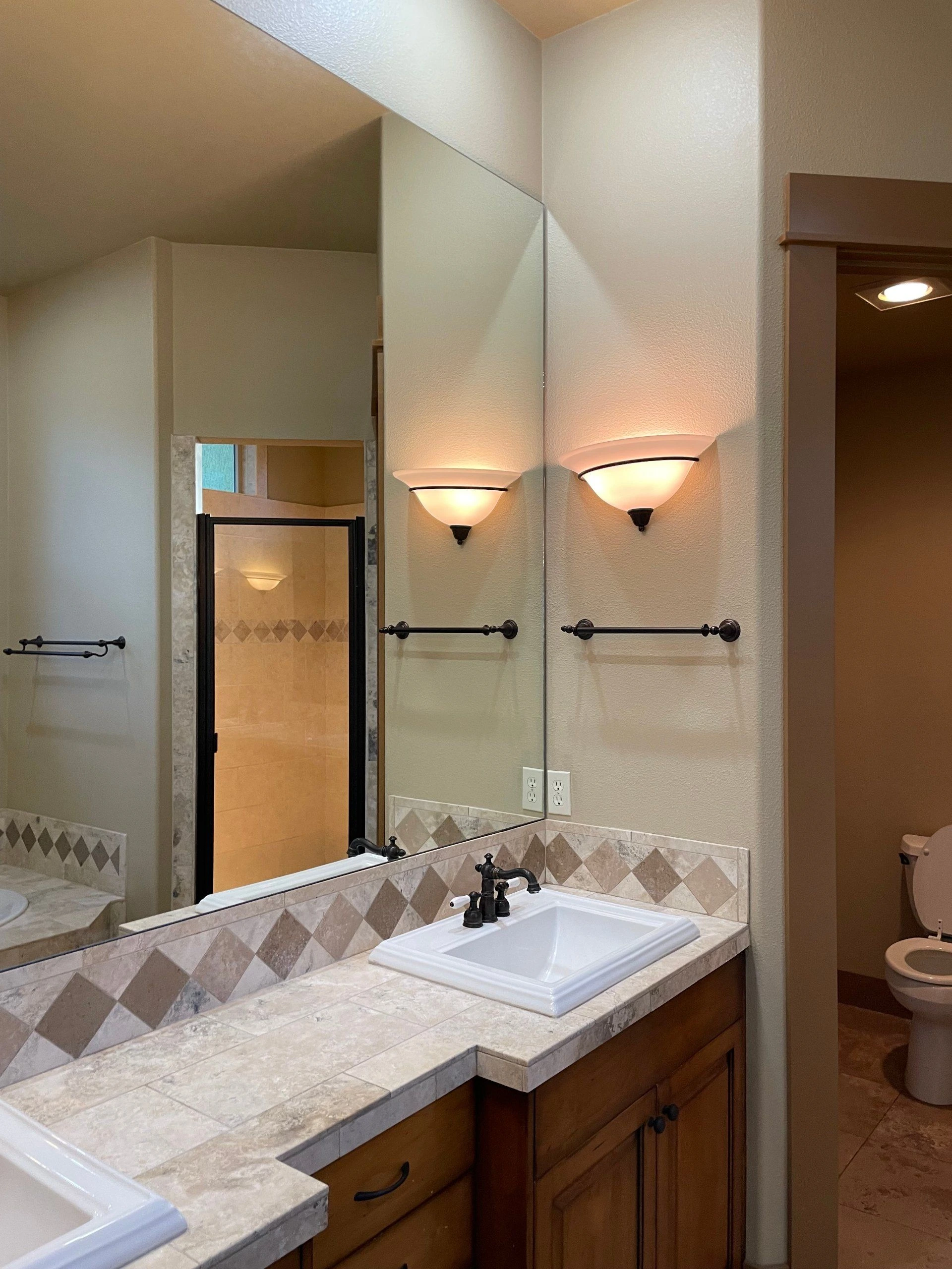 A bathroom vanity with two sinks, a large mirror, lit wall sconces, and a tiled backsplash; a shower and toilet are visible.